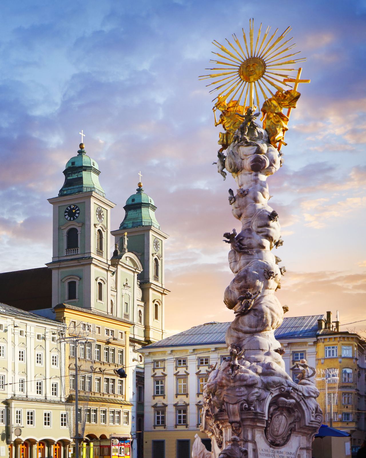Barocke Säule mit goldenem Heiligenschein im Vordergrund, umgeben von historischen Gebäuden und einer Kirche mit zwei Türmen, bei blauem Himmel mit vereinzelten Wolken im Abendlicht.