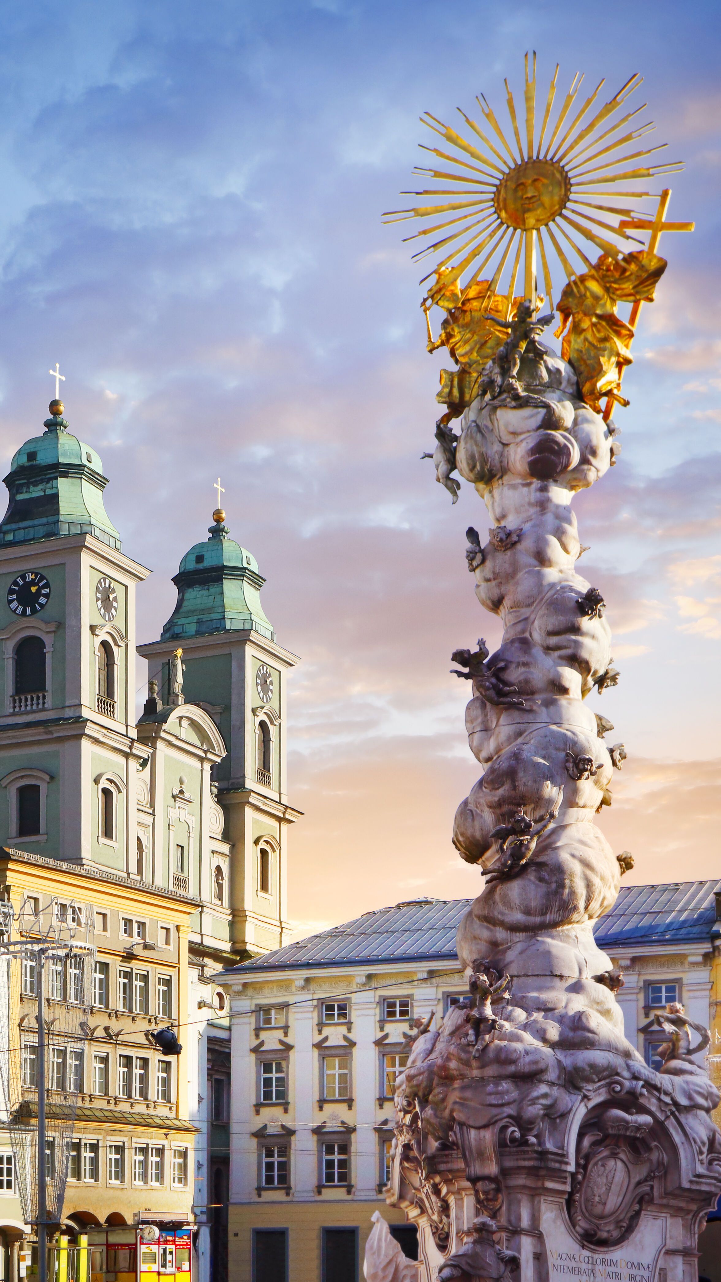 Barocke Säule mit goldenem Heiligenschein im Vordergrund, umgeben von historischen Gebäuden und einer Kirche mit zwei Türmen, bei blauem Himmel mit vereinzelten Wolken im Abendlicht.