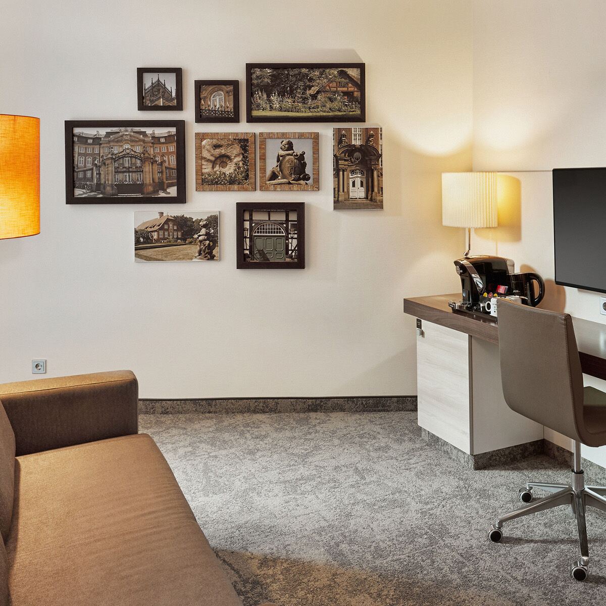 Gemütlicher Hotelzimmerbereich mit braunem Sofa, warmem Stehlampenlicht, einer Wand mit gerahmten Kunstfotos und einem Schreibtisch mit Stuhl, Schreibtischlampe und Kaffeezubehör für entspanntes Arbeiten oder Ausruhen.