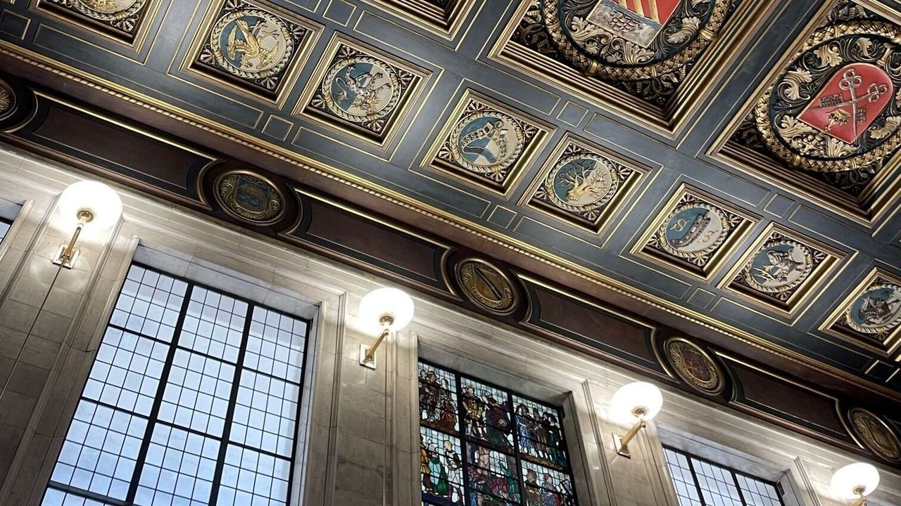 Decke mit kunstvollen, farbigen Wappenverzierungen und umgebender goldener Ornamentik über großen Fensterfronten mit buntem Bleiglas in einem eleganten, historischen Raum.
