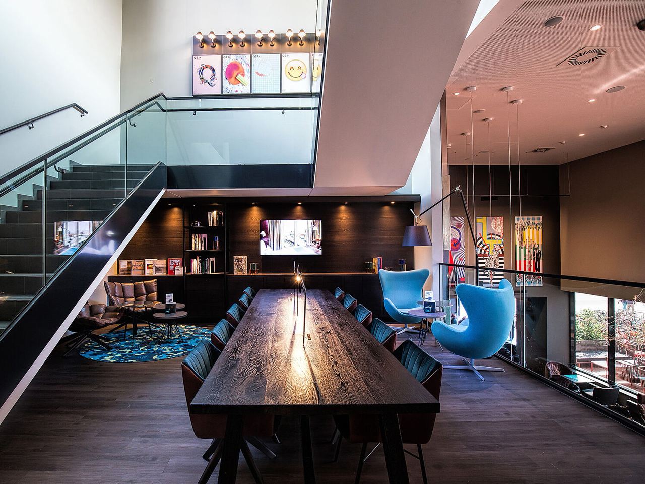 Motel One Gemeinschaftsbereich mit langem Holztisch, gemütlichen Sesseln und großen Fenstern, viel Tageslicht und stilvoller Beleuchtung