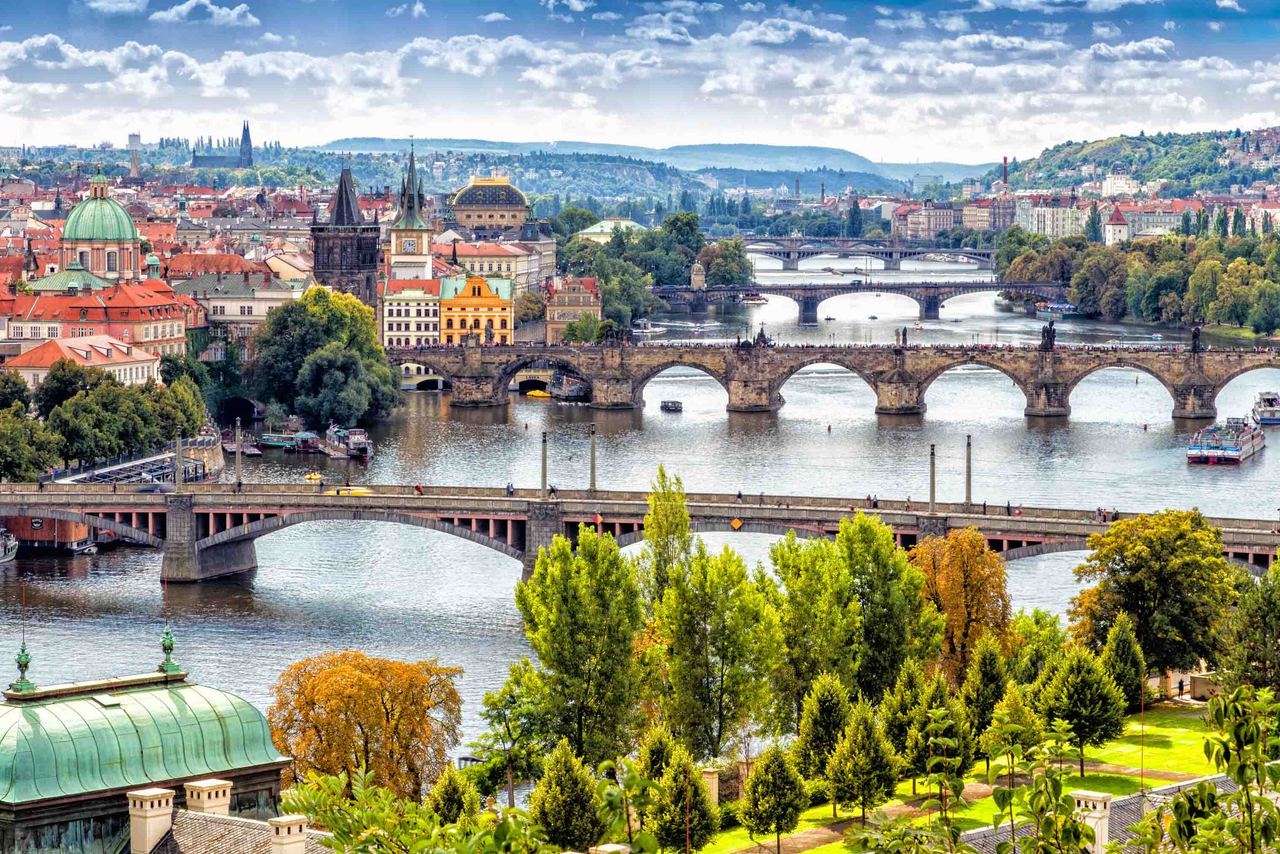 Blick auf die historische Stadt Prag mit mehreren Brücken über die Moldau, grünen Parks am Flussufer und markanten Gebäuden unter einem lebhaften Himmel.