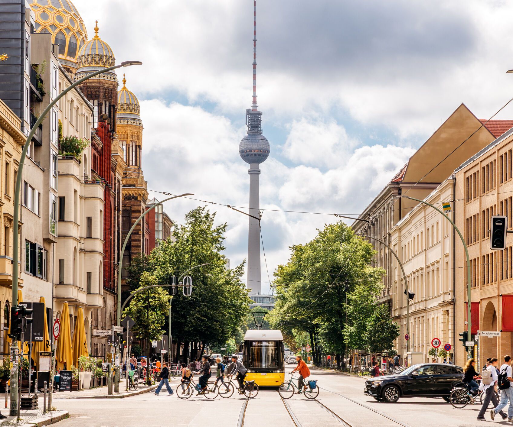 Blick auf eine lebhafte Berliner Straße mit Fußgängern, Radfahrern und einer Straßenbahn, im Hintergrund der Fernsehturm und die goldene Kuppel der Synagoge, umgeben von historischen und modernen Gebäuden an einem bewölkten Tag.