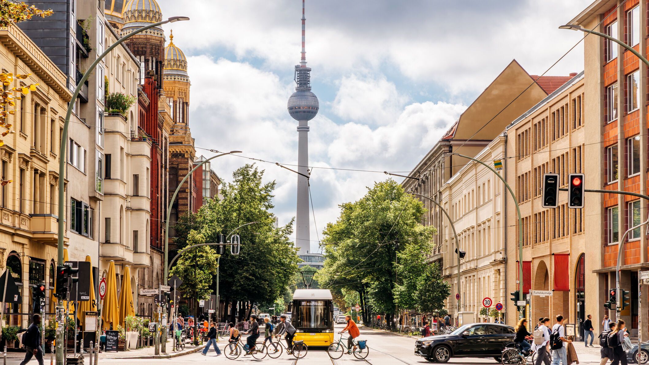 Blick auf eine lebhafte Berliner Straße mit Fußgängern, Radfahrern und einer Straßenbahn, im Hintergrund der Fernsehturm und die goldene Kuppel der Synagoge, umgeben von historischen und modernen Gebäuden an einem bewölkten Tag.