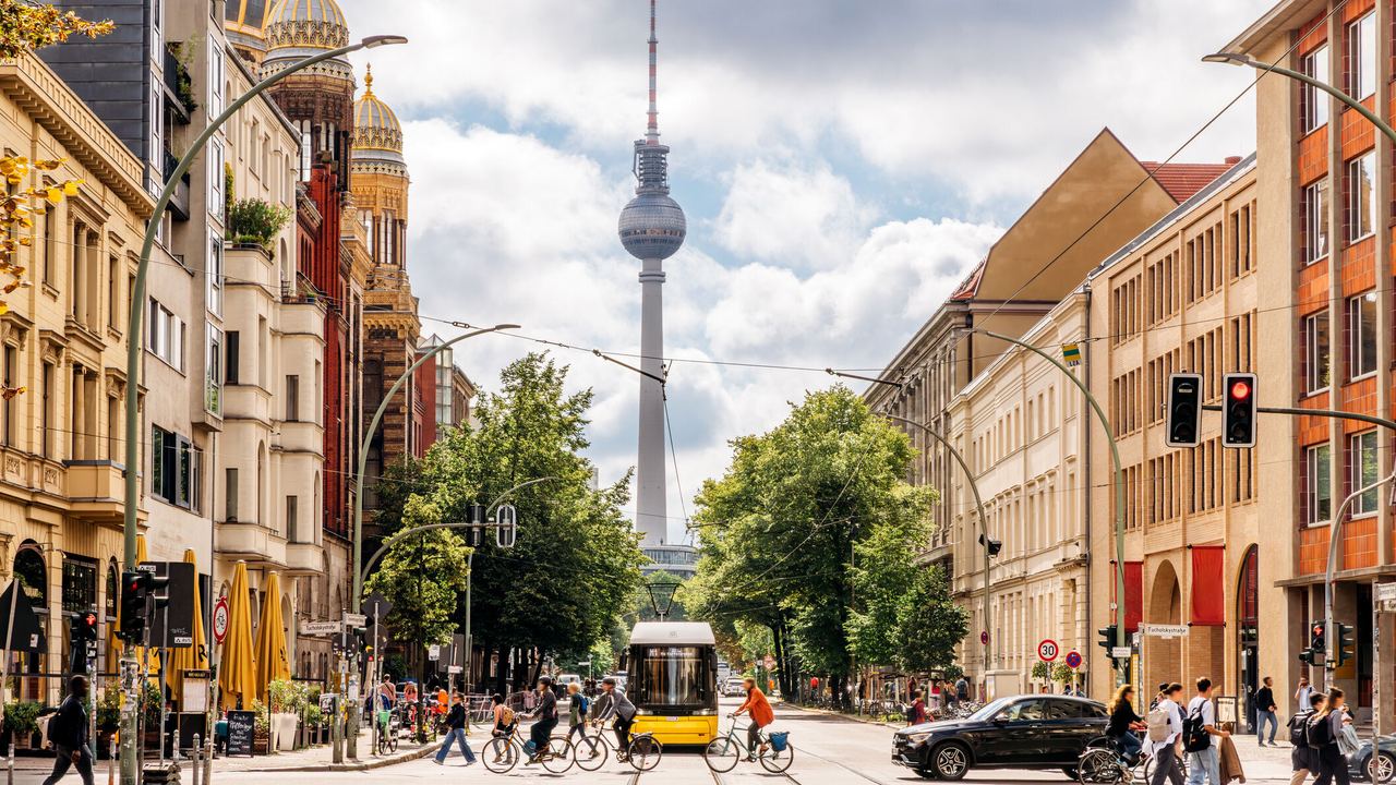Blick auf eine lebhafte Berliner Straße mit Fußgängern, Radfahrern und einer Straßenbahn, im Hintergrund der Fernsehturm und die goldene Kuppel der Synagoge, umgeben von historischen und modernen Gebäuden an einem bewölkten Tag.