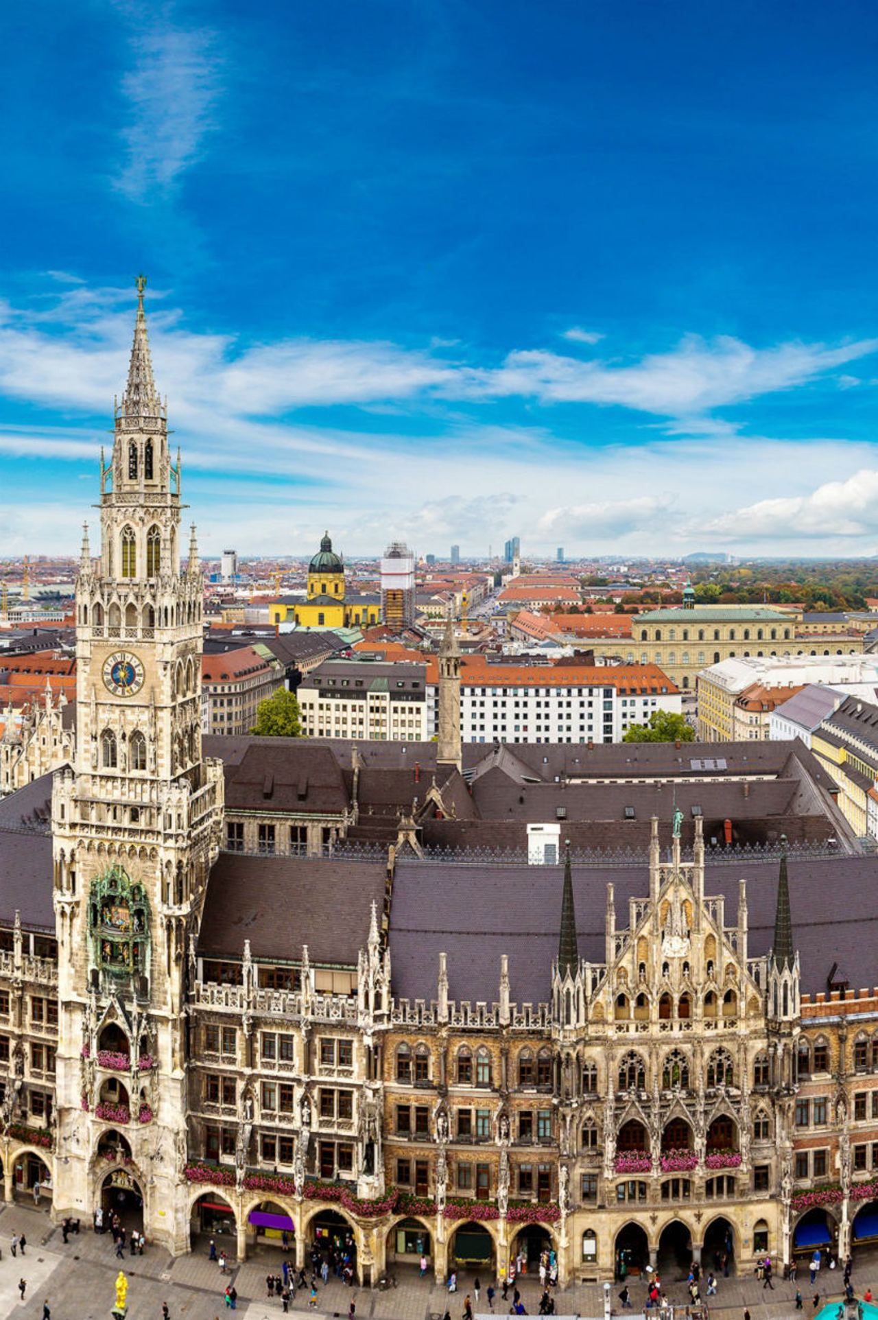 Blick auf den belebten Marienplatz in München mit dem historischen Neuen Rathaus und seinem markanten Glockenspiel unter blauem Himmel. Menschen bewegen sich auf dem Platz, der von altstädtischer Architektur umgeben ist.