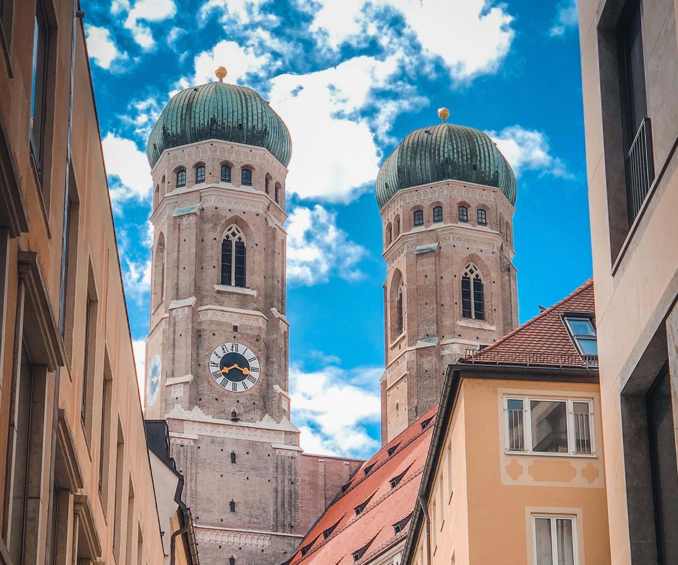 Blick auf die markanten Zwillingstürme der Frauenkirche in München, eingerahmt von engen Straßenzügen und blauen Himmel mit Wolken.