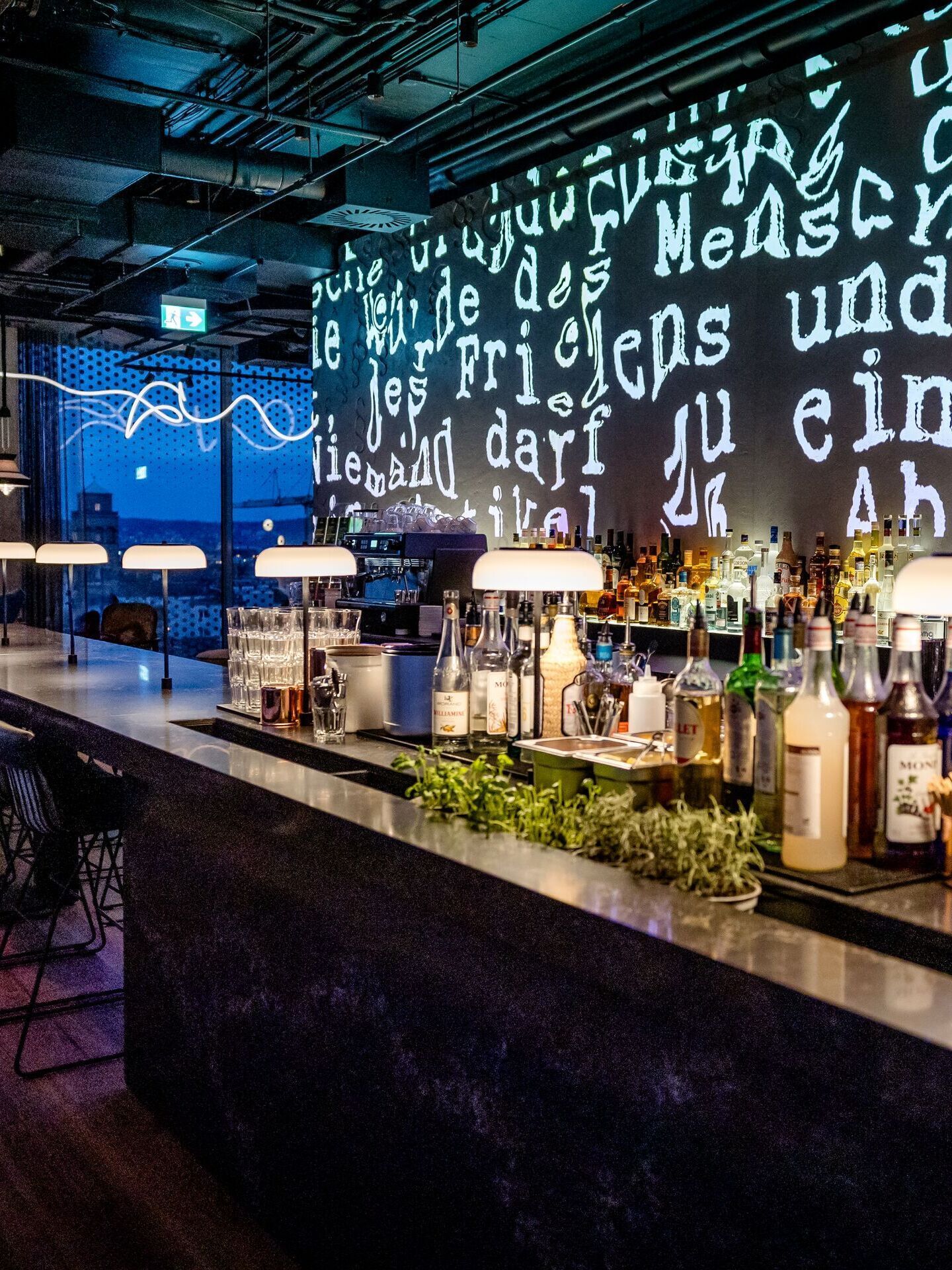 Moderne Hotelbar mit beleuchtetem Tresen, zahlreichen Spirituosen und eleganter, stimmungsvoller Atmosphäre. Große Fenster bieten Blick auf die Stadt bei Dämmerung. Kreative Wandprojektion mit Text verleiht dem Raum einen urbanen, künstlerischen Charakter.