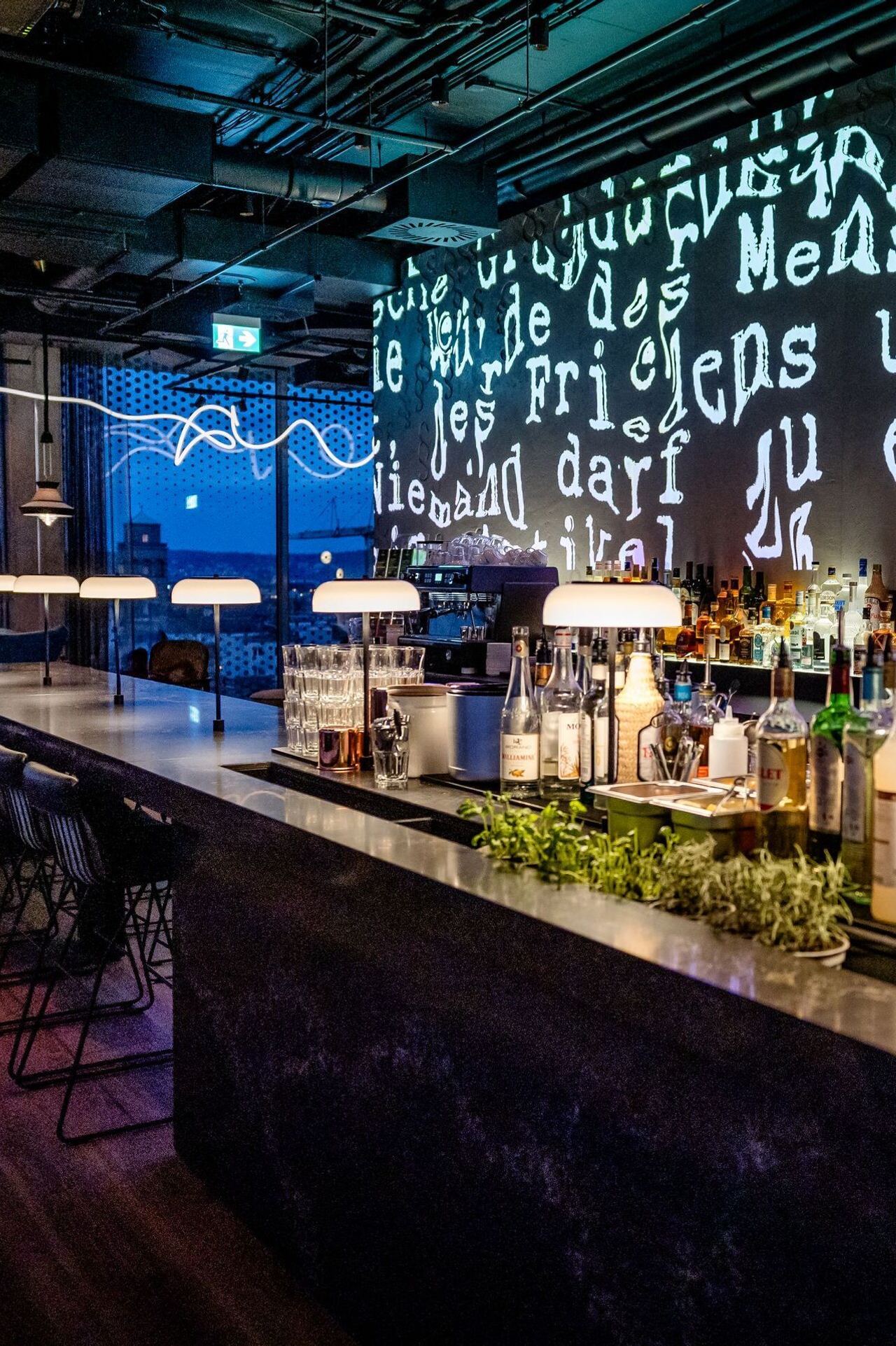 Moderne Hotelbar mit beleuchtetem Tresen, zahlreichen Spirituosen und eleganter, stimmungsvoller Atmosphäre. Große Fenster bieten Blick auf die Stadt bei Dämmerung. Kreative Wandprojektion mit Text verleiht dem Raum einen urbanen, künstlerischen Charakter.