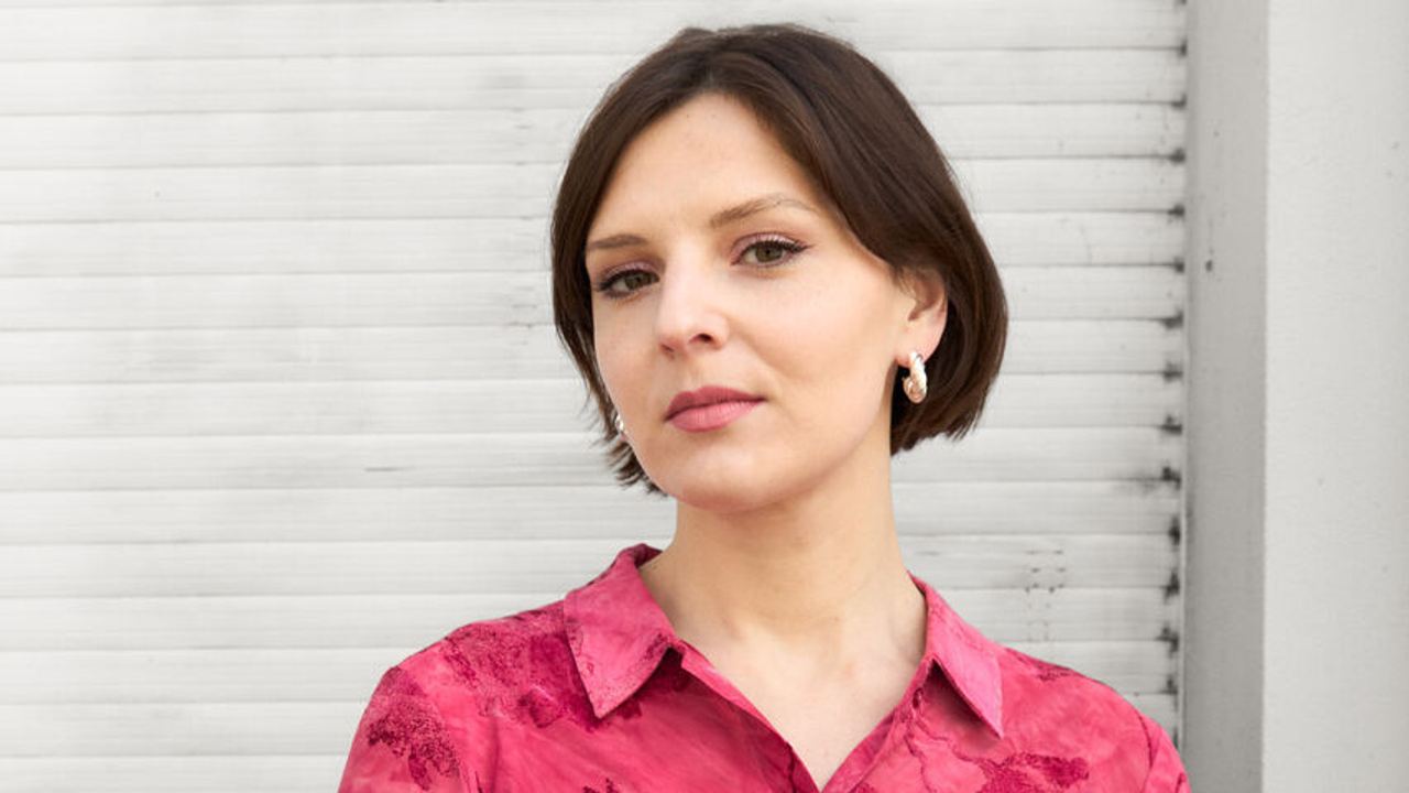 Porträt einer selbstbewusst wirkenden Frau in rosa geblümter Bluse vor hellem Hintergrund, die Ruhe und Gelassenheit ausstrahlt.