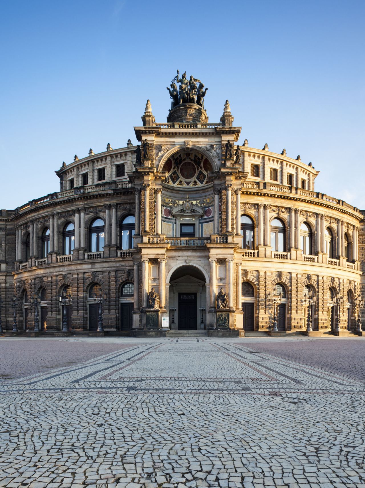 Prächtiges historisches Gebäude mit aufwändiger Fassade und großer Skulptur auf dem Dach, umgeben von offenem, gepflastertem Platz unter klarem Himmel.