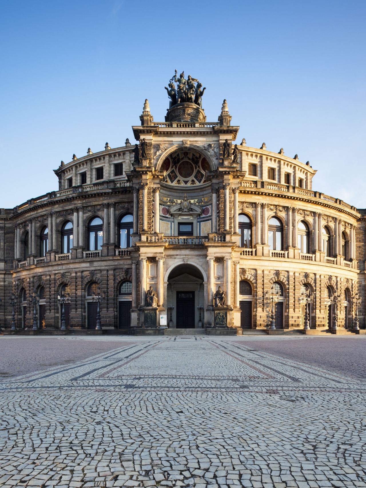 Prächtiges historisches Gebäude mit aufwändiger Fassade und großer Skulptur auf dem Dach, umgeben von offenem, gepflastertem Platz unter klarem Himmel. Prächtiges historisches Gebäude mit aufwändiger Fassade und großer Skulptur auf dem Dach, umgeben von offenem, gepflastertem Platz unter klarem Himmel.