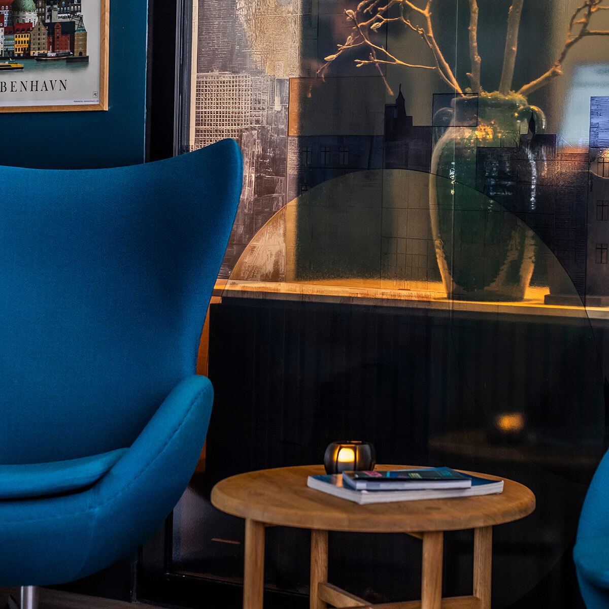 Gemütliche Sitzecke mit modernem, blauem Designstuhl, kleinem Holztisch mit Büchern, stilvoll dekoriert mit Vase und Zweigen, neben einer Treppe und einem farbenfrohen Stadtposter von Kopenhagen.
