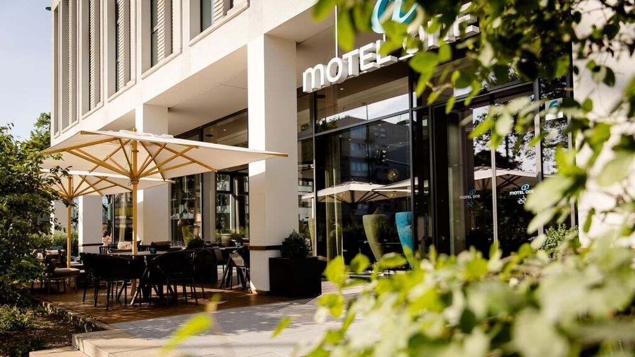 Außenansicht eines Hotels mit Terrasse, die mit Sonnenschirmen und Tischen zum entspannten Verweilen einlädt. Grüne Pflanzen umrahmen den Eingangsbereich.