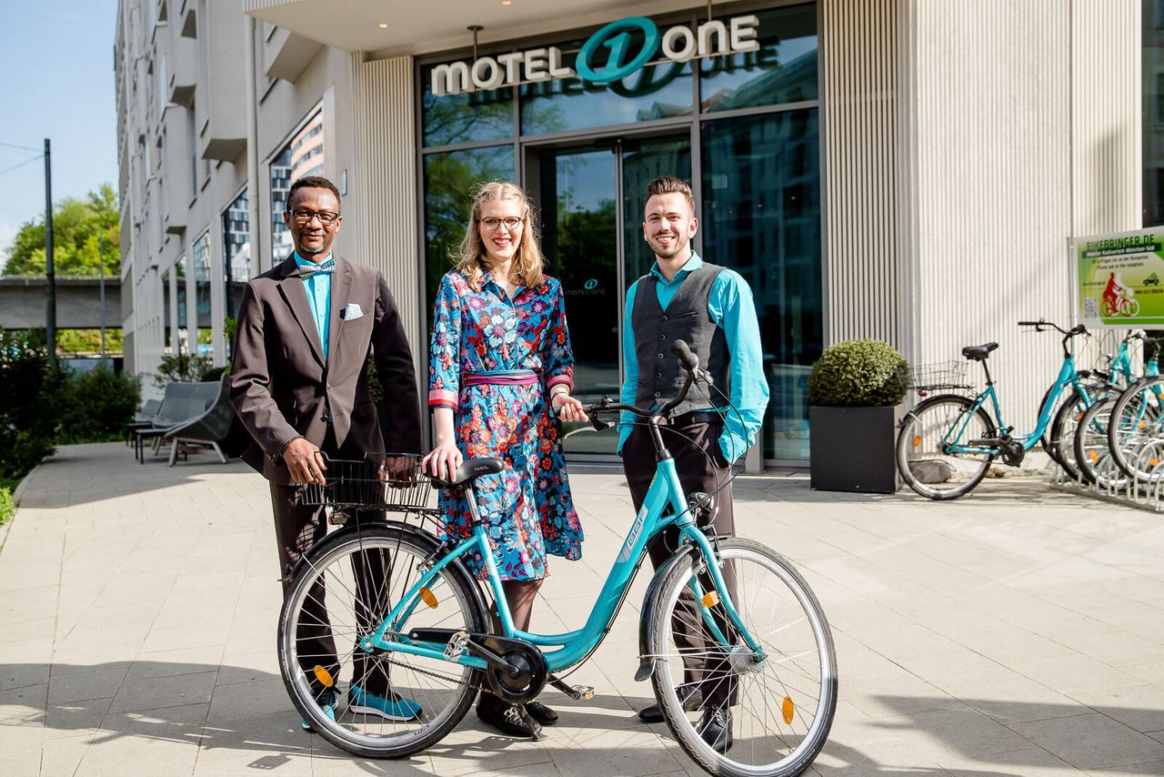 Drei freundliche Hotelmitarbeiter stehen vor dem Eingang des Motels, zwei Männer im eleganten Outfit, eine Frau in buntem Kleid, neben einem modernen blauen Fahrrad.