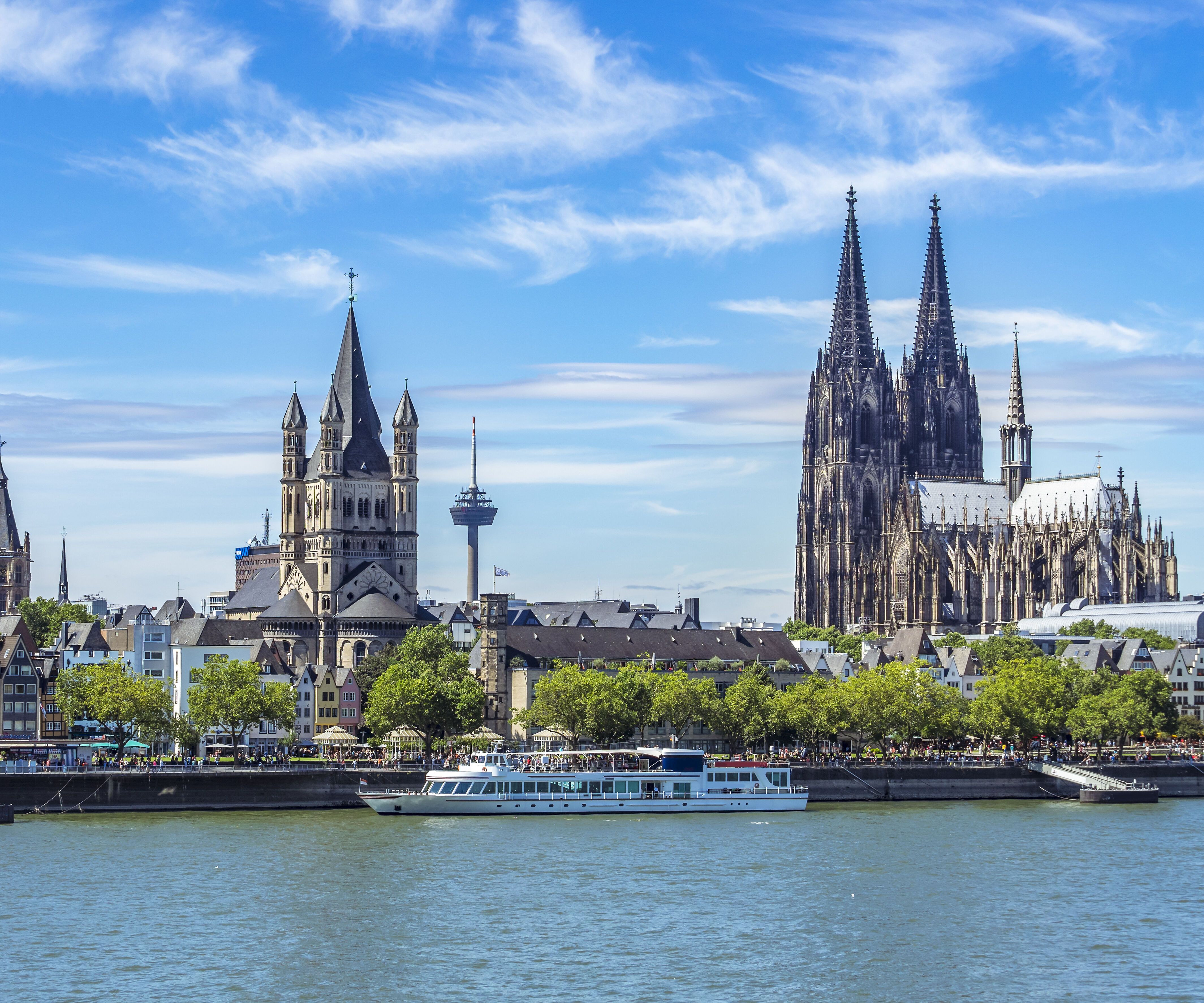 Panoramablick auf den Rhein in Köln mit markanten Kirchen, darunter der Kölner Dom, und grünen Uferbereichen bei blauem Himmel.