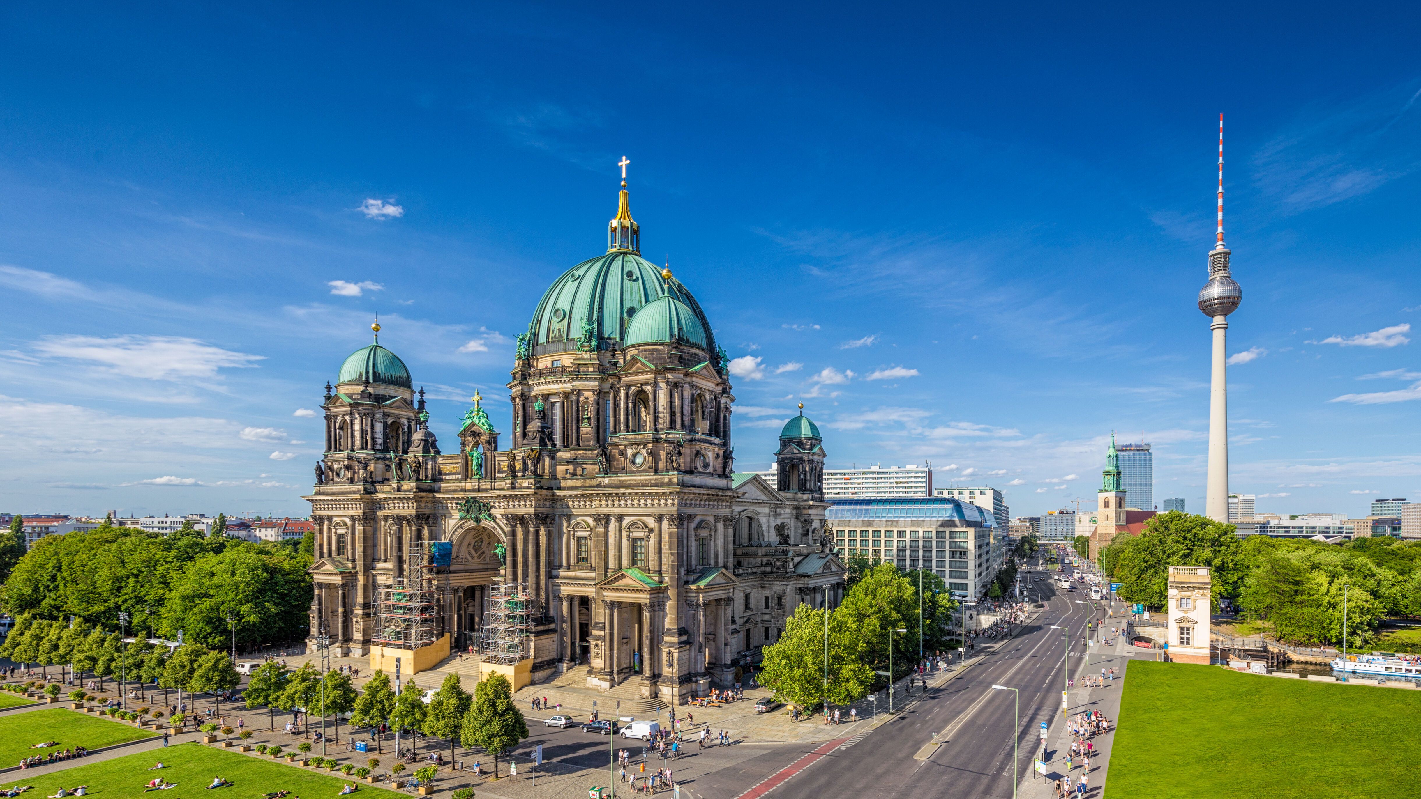 Berliner Dom mit grünem Kupferdach und Kuppeln, umgeben von Parks mit liegenden Menschen. Im Hintergrund ist der Fernsehturm sichtbar, ein Symbol Berlins, bei klarem blauem Himmel. Die Straße vor dem Dom ist belebt mit Fußgängern und Fahrzeugen.