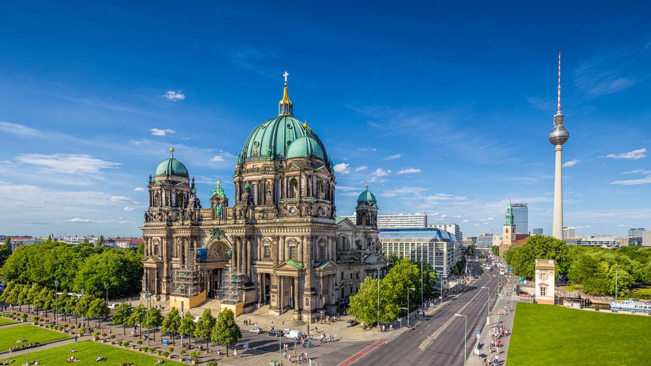 Berliner Dom mit grünem Kupferdach und Kuppeln, umgeben von Parks mit liegenden Menschen. Im Hintergrund ist der Fernsehturm sichtbar, ein Symbol Berlins, bei klarem blauem Himmel. Die Straße vor dem Dom ist belebt mit Fußgängern und Fahrzeugen.