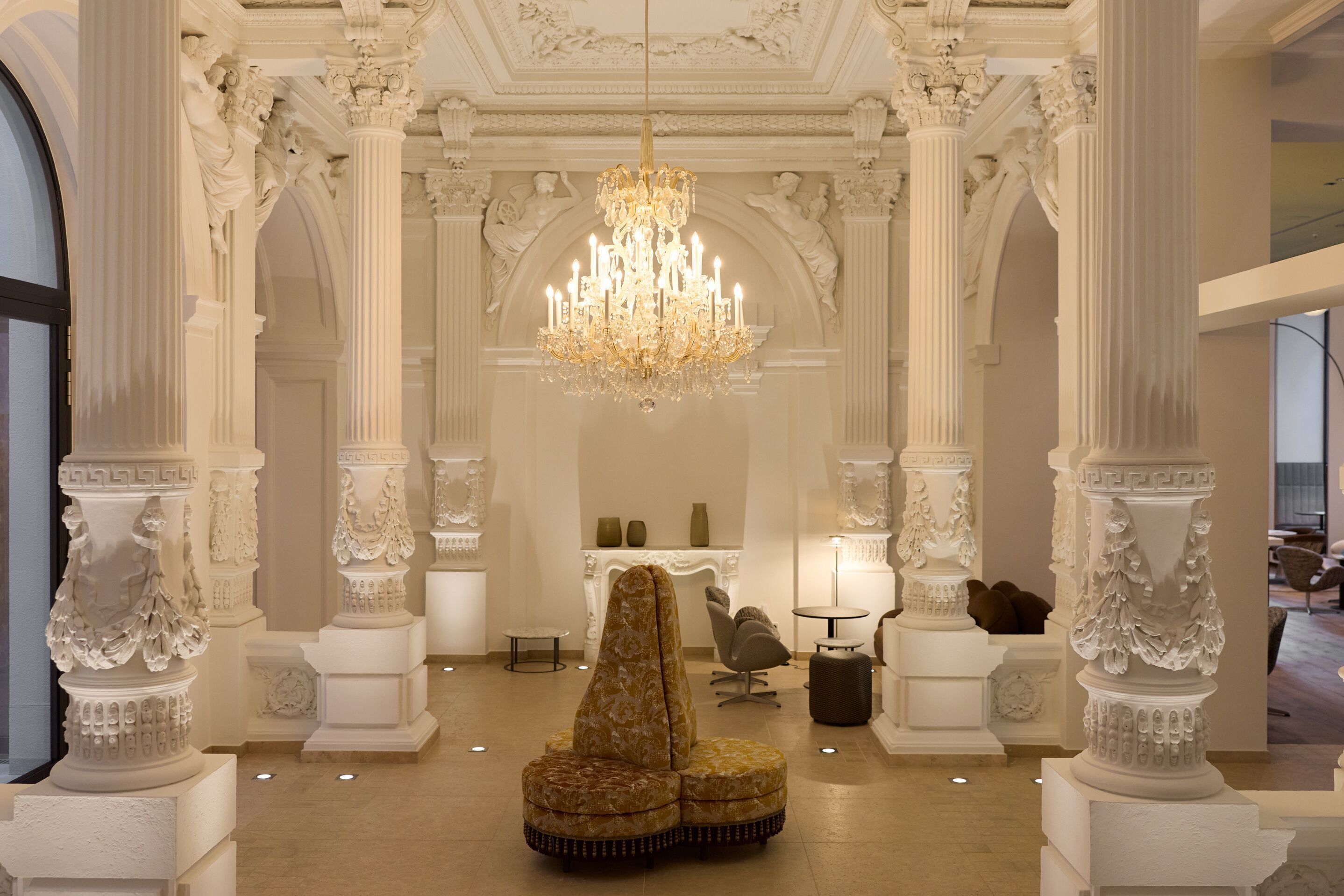 Helle Lobby mit prunkvollen Stuckdecken, sechs kunstvoll verzierten Säulen, elegantem Kronleuchter und bequemen Sitzgelegenheiten, die eine luxuriöse und einladende Atmosphäre schaffen.