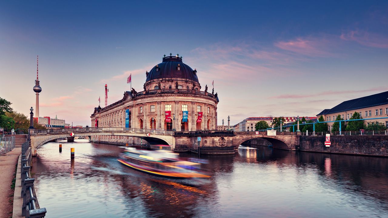 Das historische Bode-Museum in Berlin am Abend, mit sanfter Beleuchtung, ruhigem Fluss im Vordergrund und einem vorbeifahrenden Boot, das Bewegung und lebendige Atmosphäre vermittelt.