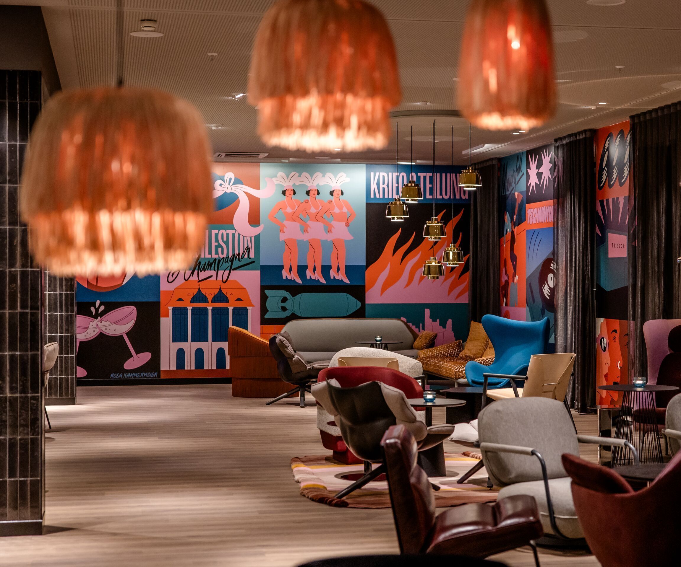 Hotel Berlin Potsdamer Platz Motel One moderne Lounge mit bunten, künstlerischen Wandbildern, stilvollen Sesseln und Sofas in warmem Licht, ideal zum Entspannen und geselligen Beisammensein.