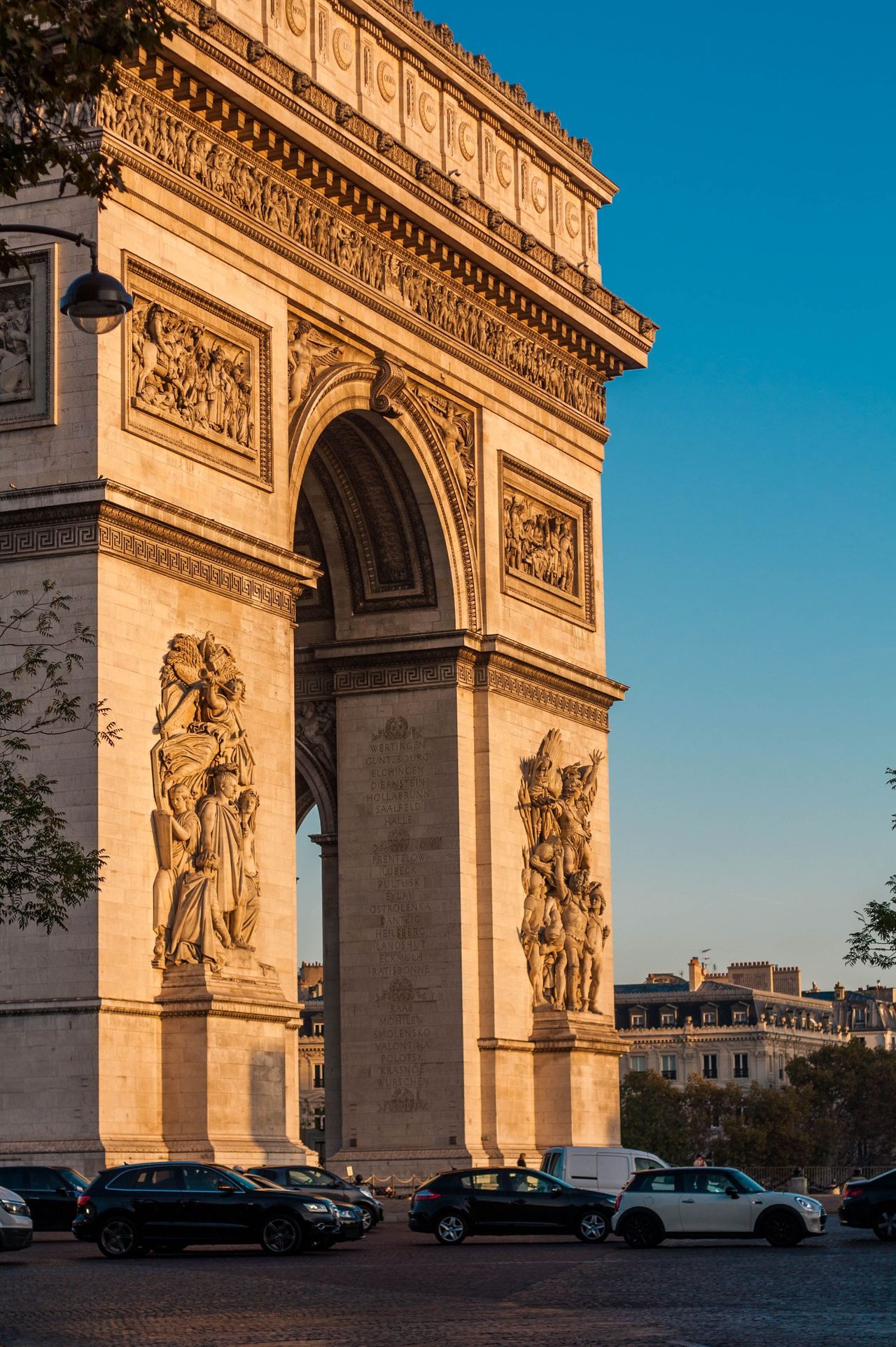 Der Arc de Triomphe in Paris bei Sonnenuntergang, mit detailreichen Reliefs und Skulpturen an den Pfeilern, davor parkende Autos und klare, blaue Himmel.