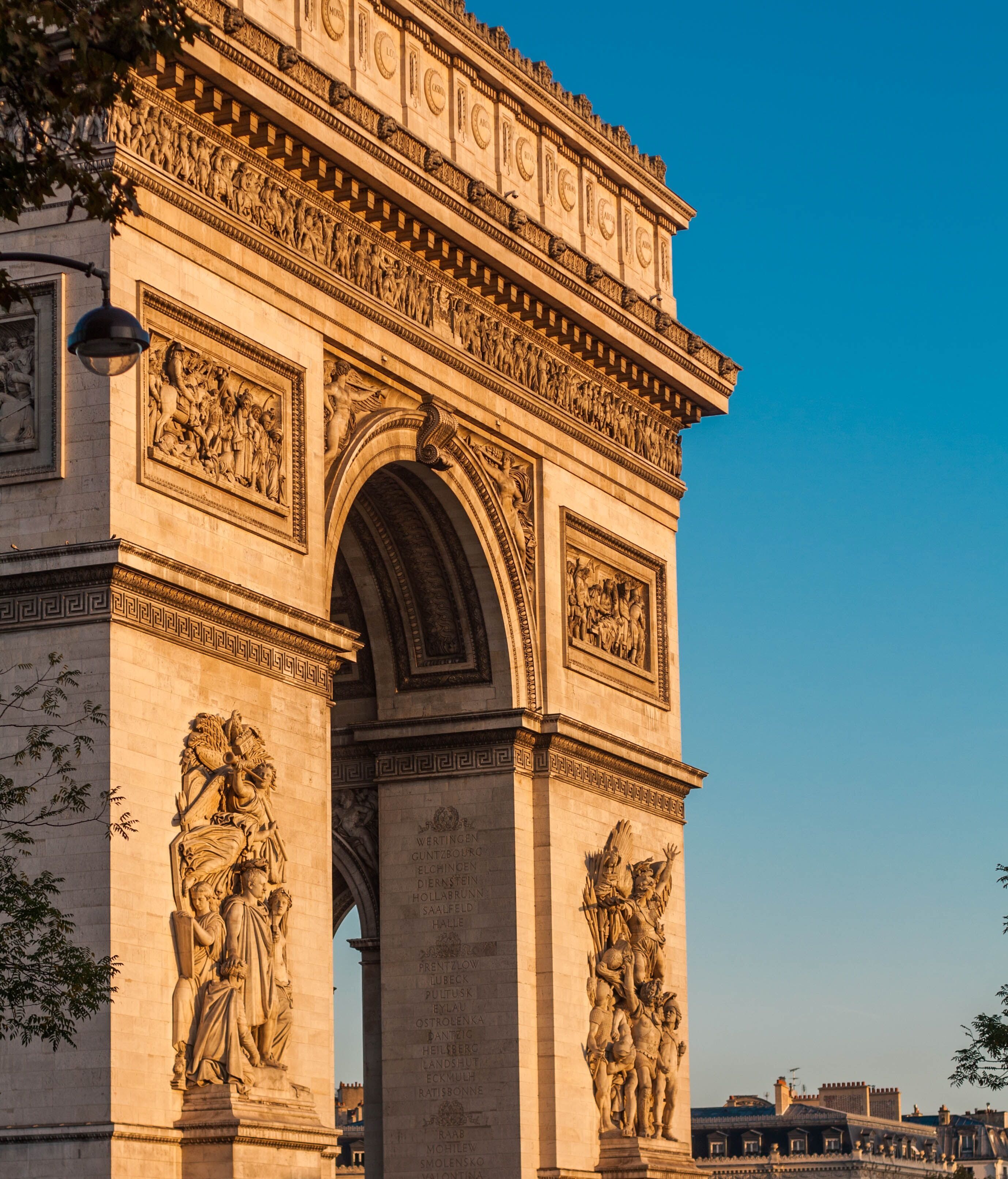 Der Arc de Triomphe in Paris bei Sonnenuntergang, mit detailreichen Reliefs und Skulpturen an den Pfeilern, davor parkende Autos und klare, blaue Himmel.