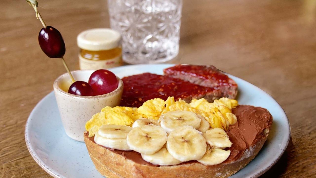 Frühstücksteller mit einem Toast, bestrichen mit Schokolade, belegt mit Bananenscheiben, dazu Rührei, eine Scheibe Wurst und ein kleiner Becher mit Kirschen auf einem Holztisch.