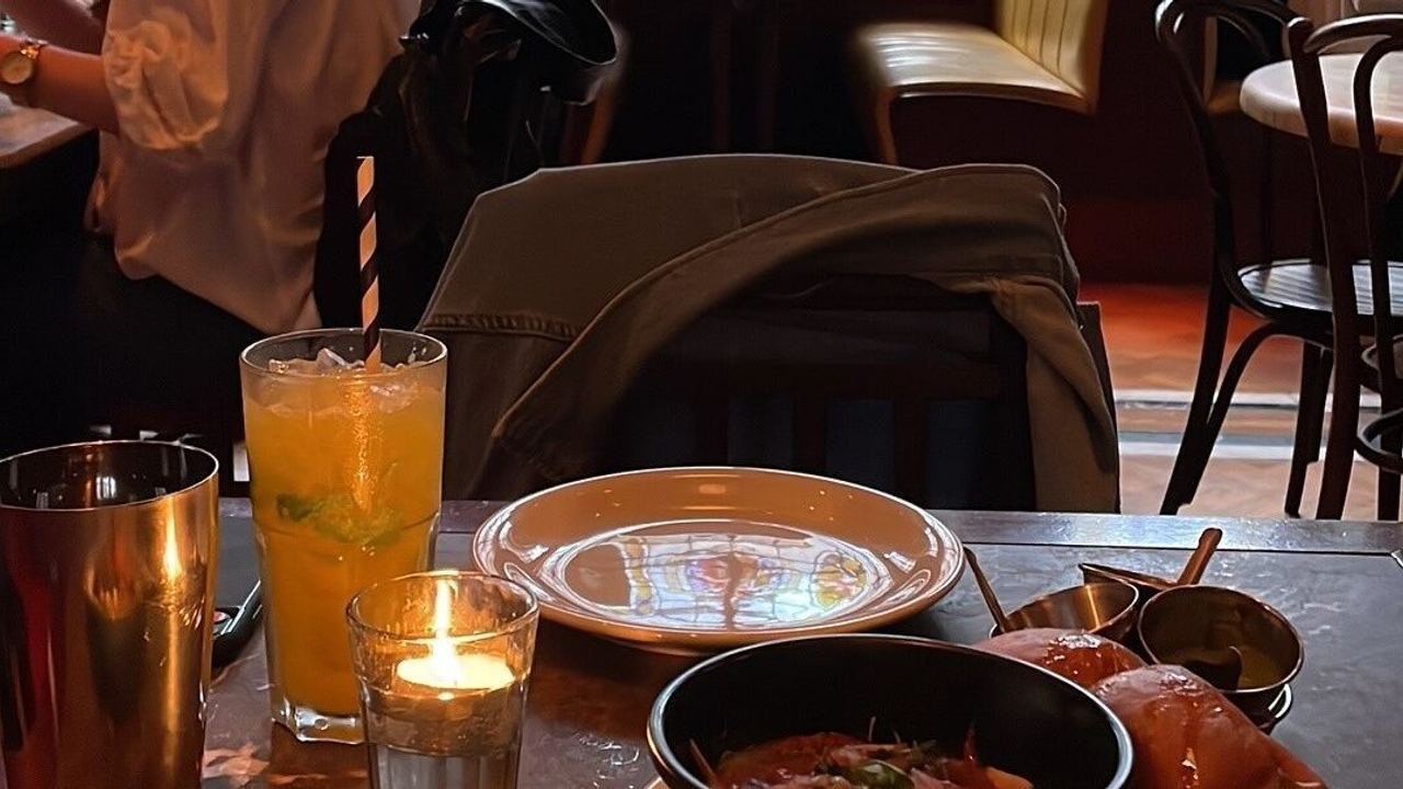 Restaurant mit bunten Buntglasfenstern. Auf dem Tisch stehen ein Glas mit orangefarbenem Getränk, eine Kerze und ein Teller mit einer Mahlzeit.
