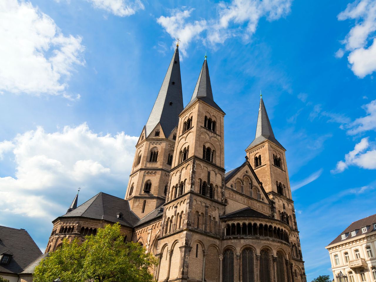 Hotel Bonn dreiturmige, historische Kirche mit spitzen Dächern aus hellem Sandstein vor blauem Himmel, umgeben von Gebäuden und Bäumen.