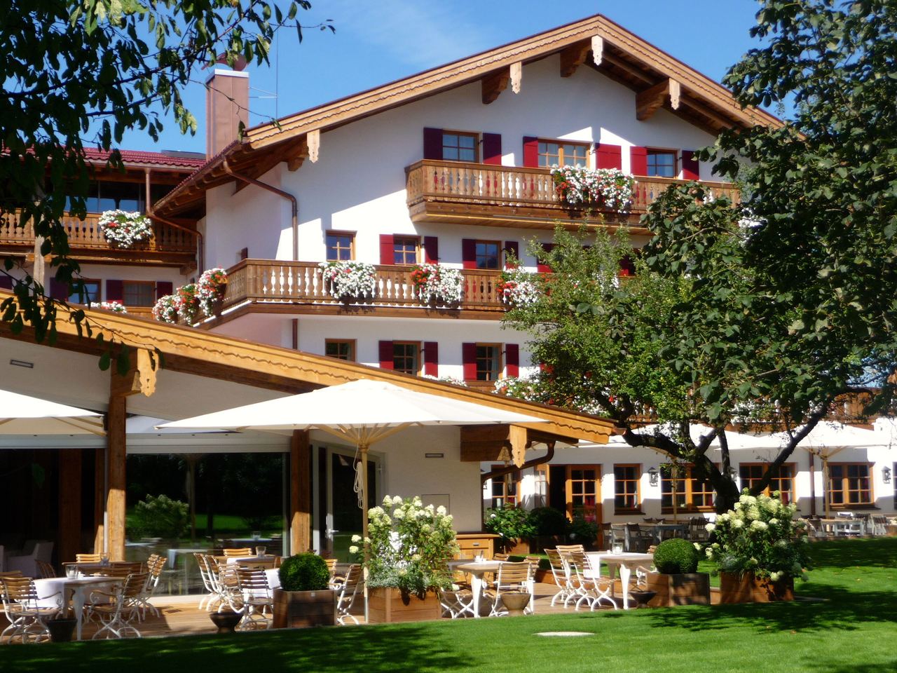 Traditionelles Hotelgebäude mit Holzbalkonen und blühenden Pflanzen, gemütliche Außenterrasse mit Sitzgelegenheiten, umgeben von grünem Garten und Bäumen, einladende Atmosphäre für Gäste zum Entspannen und Genießen.