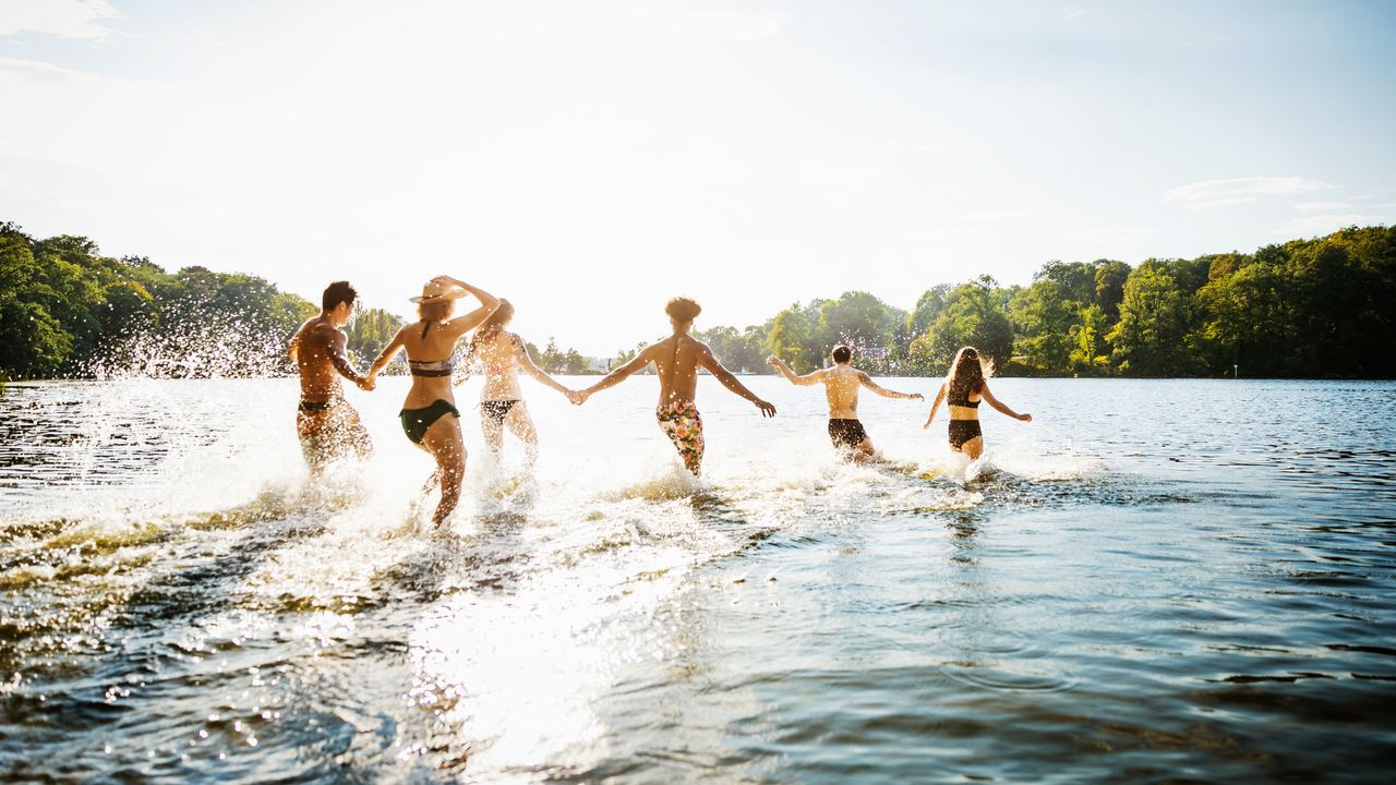 Sechs junge Menschen laufen lachend und handhaltend in einem See, umgeben von sommerlicher Natur, unter blauem Himmel.