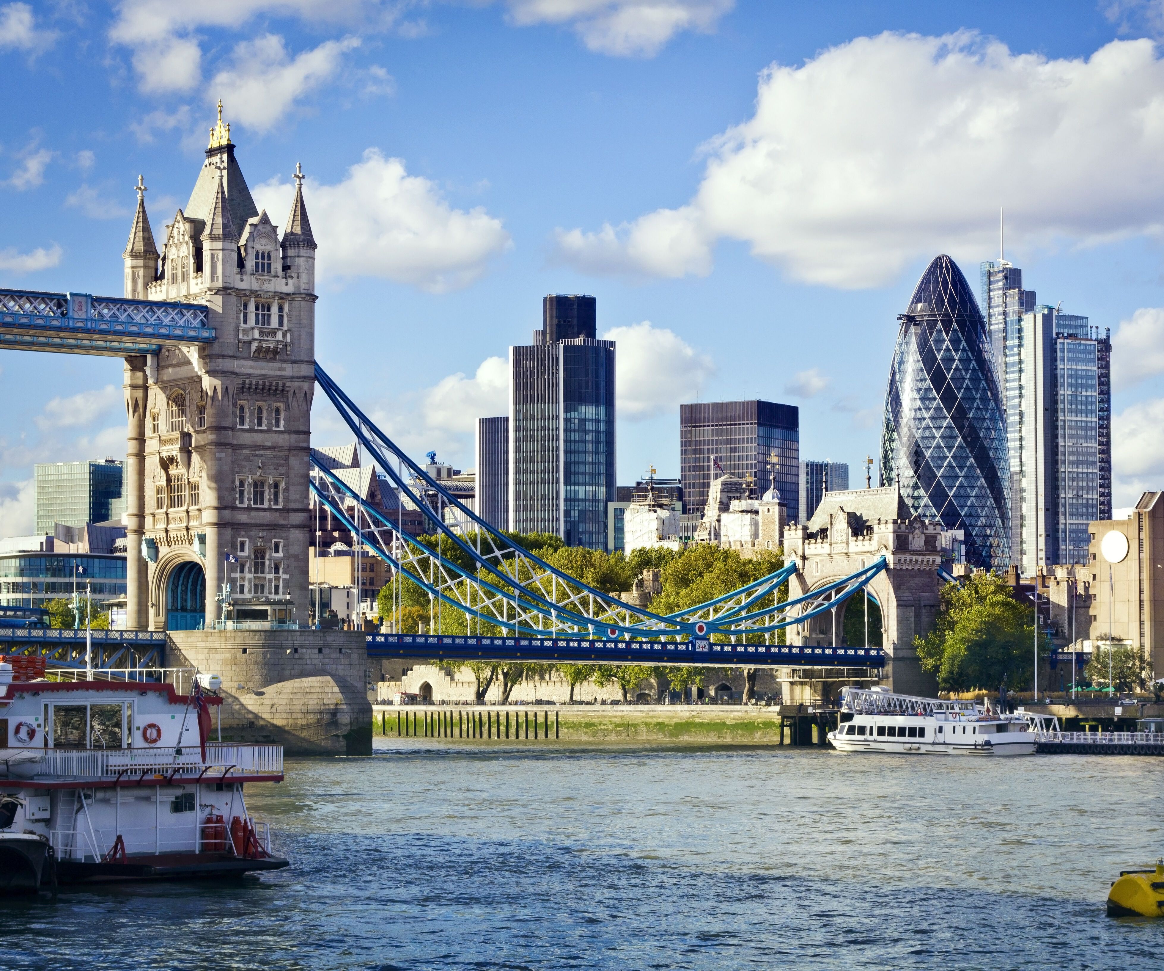 Blick auf die Tower Bridge in London mit moderner Skyline im Hintergrund, darunter das markante Gebäude „The Gherkin“. Der Fluss Themse fließt ruhig, Boote sind am Ufer zu sehen, bei klarem Himmel mit vereinzelten Wolken.