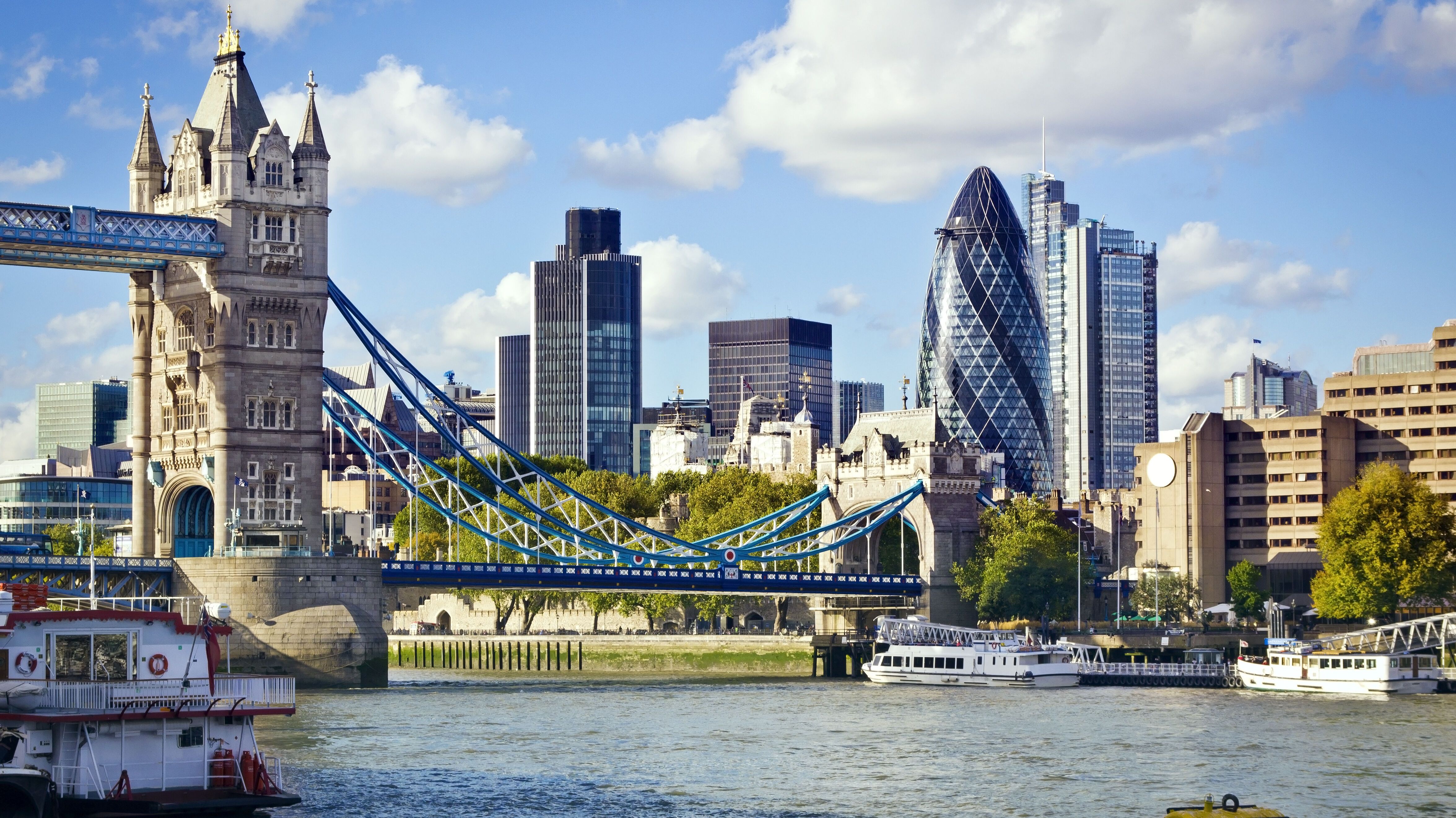 Blick auf die Tower Bridge in London mit moderner Skyline im Hintergrund, darunter das markante Gebäude „The Gherkin“. Der Fluss Themse fließt ruhig, Boote sind am Ufer zu sehen, bei klarem Himmel mit vereinzelten Wolken.
