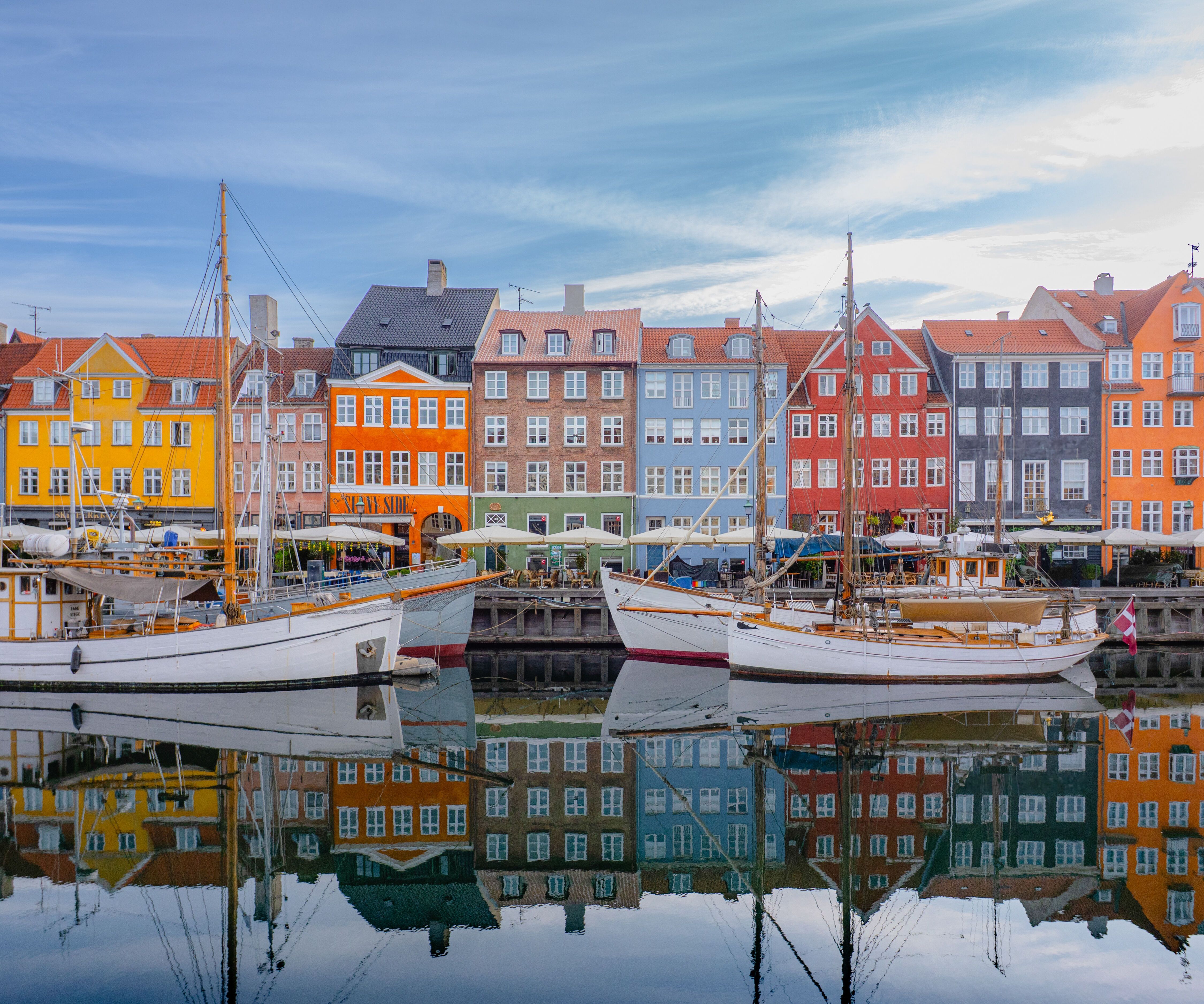 Bunte Häuserreihe am Wasser mit Segelbooten im Hafen, spiegelnd im ruhigen Wasser, vermittelt maritimes Flair und einladende Atmosphäre eines historischen Stadtviertels.