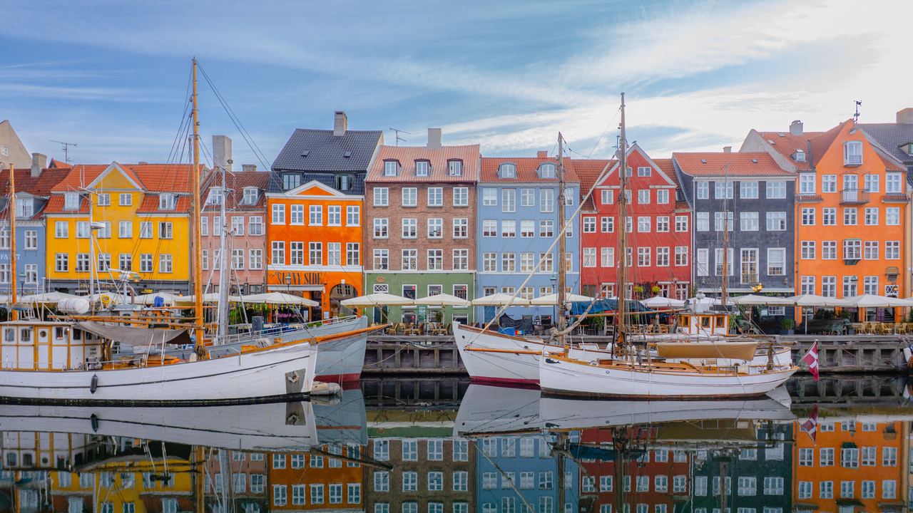 Bunte Häuserreihe am Wasser mit Segelbooten im Hafen, spiegelnd im ruhigen Wasser, vermittelt maritimes Flair und einladende Atmosphäre eines historischen Stadtviertels.