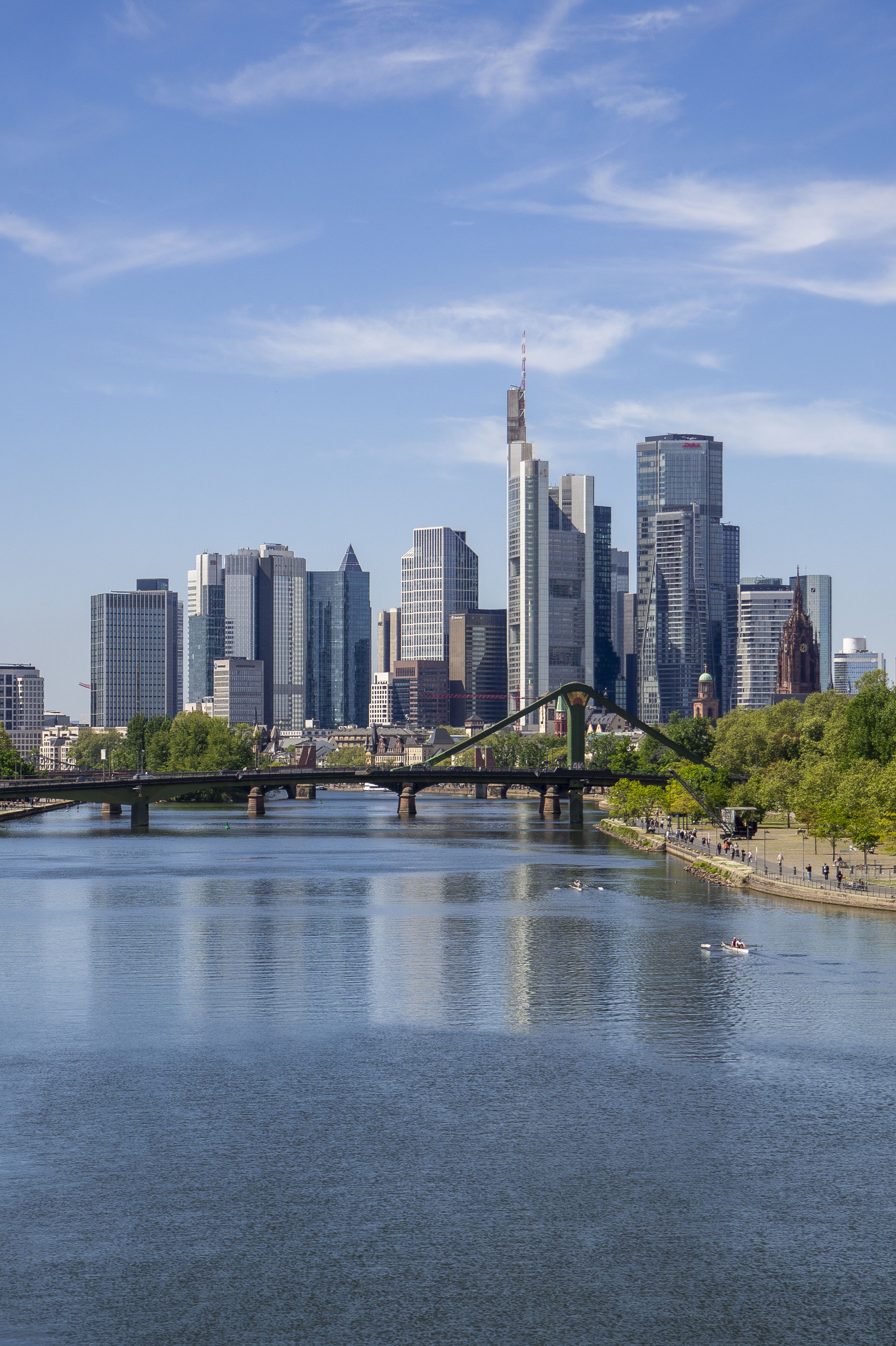 Blick auf die moderne Skyline einer Stadt mit hohen Glasgebäuden, einem Fluss im Vordergrund und einer grünen Uferpromenade bei klarem Himmel.