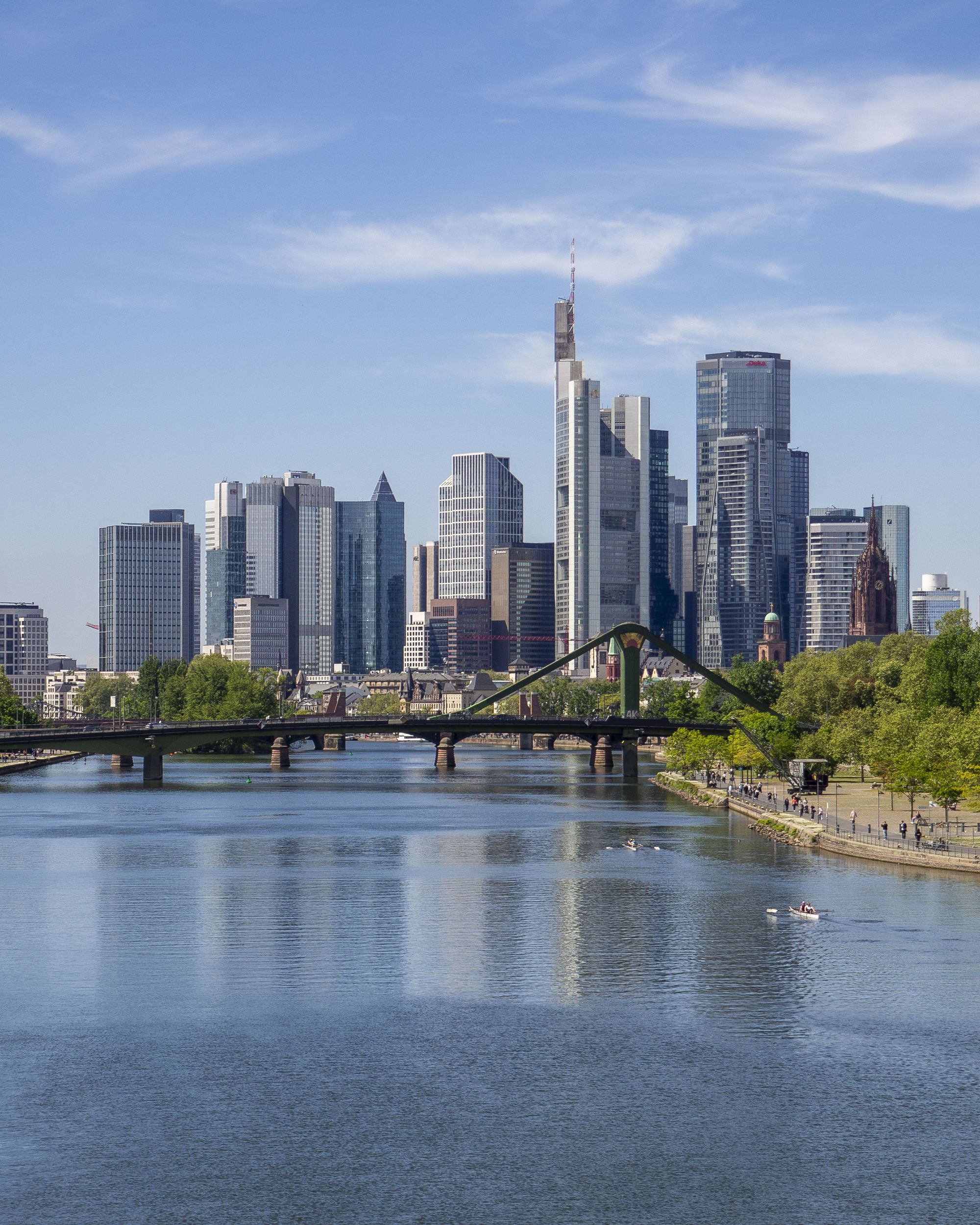 Blick auf die moderne Skyline einer Stadt mit hohen Glasgebäuden, einem Fluss im Vordergrund und einer grünen Uferpromenade bei klarem Himmel.