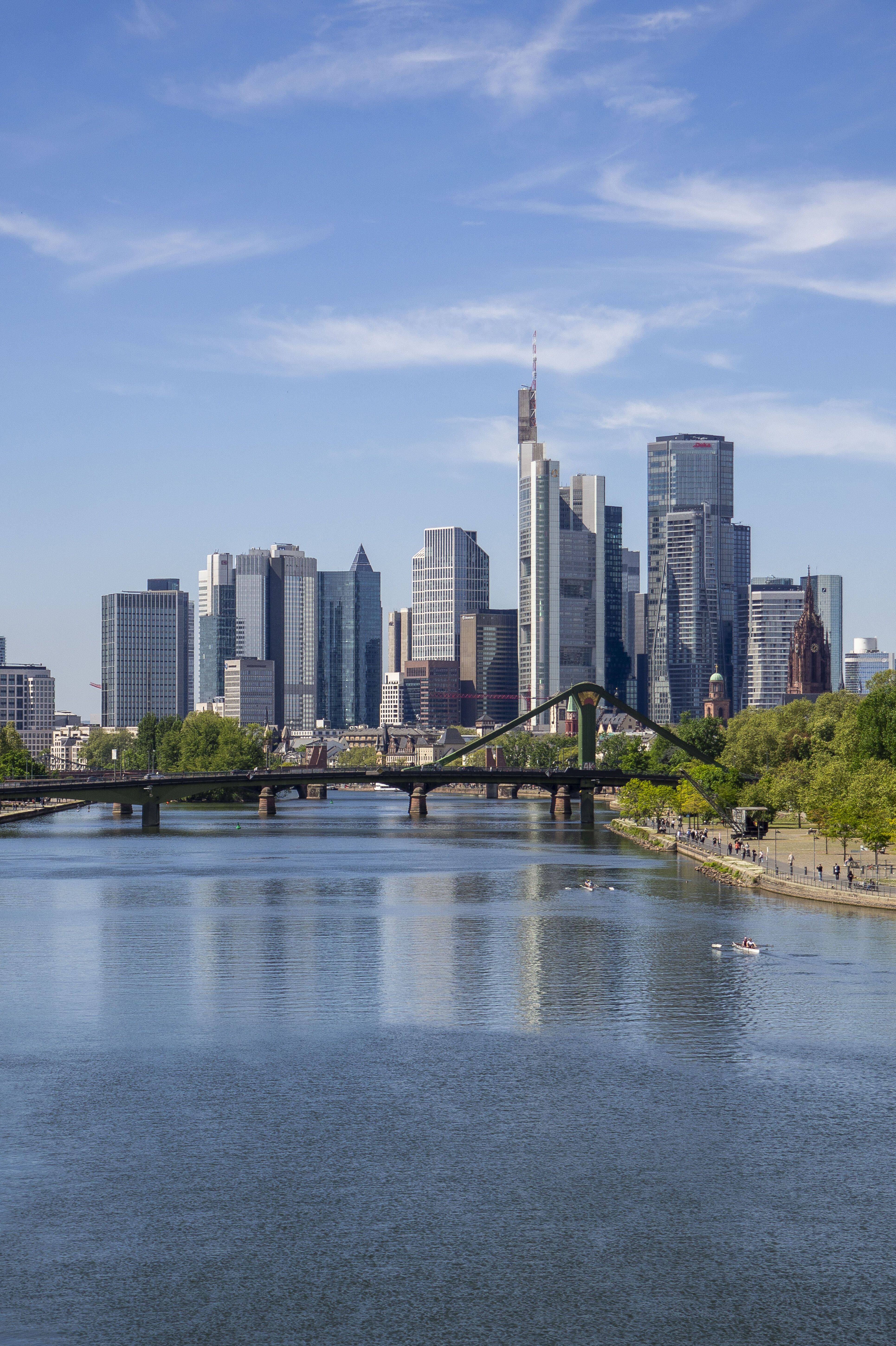 Blick auf die moderne Skyline einer Stadt mit hohen Glasgebäuden, einem Fluss im Vordergrund und einer grünen Uferpromenade bei klarem Himmel.