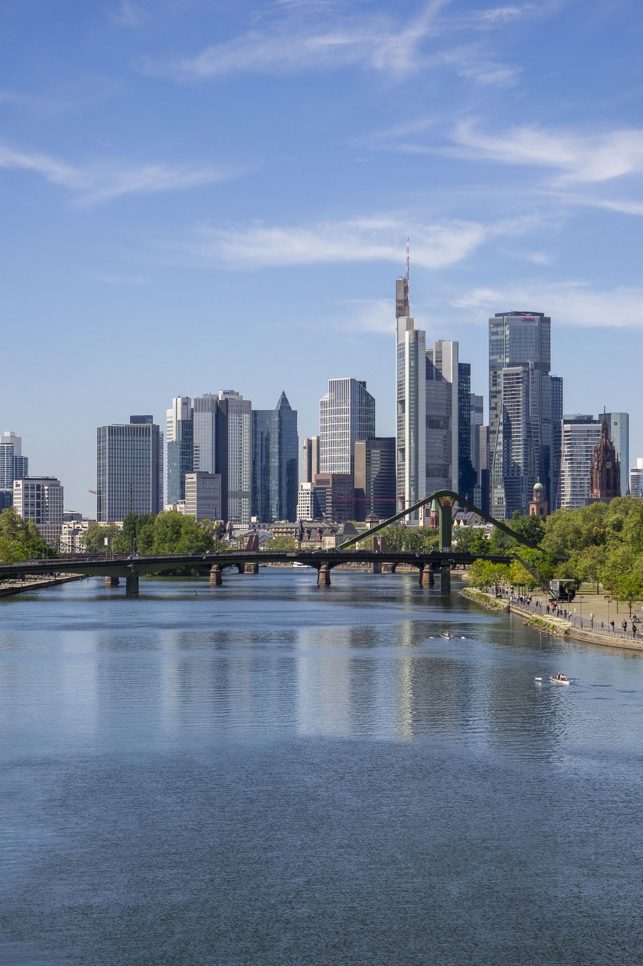 Blick auf die moderne Skyline einer Stadt mit hohen Glasgebäuden, einem Fluss im Vordergrund und einer grünen Uferpromenade bei klarem Himmel.