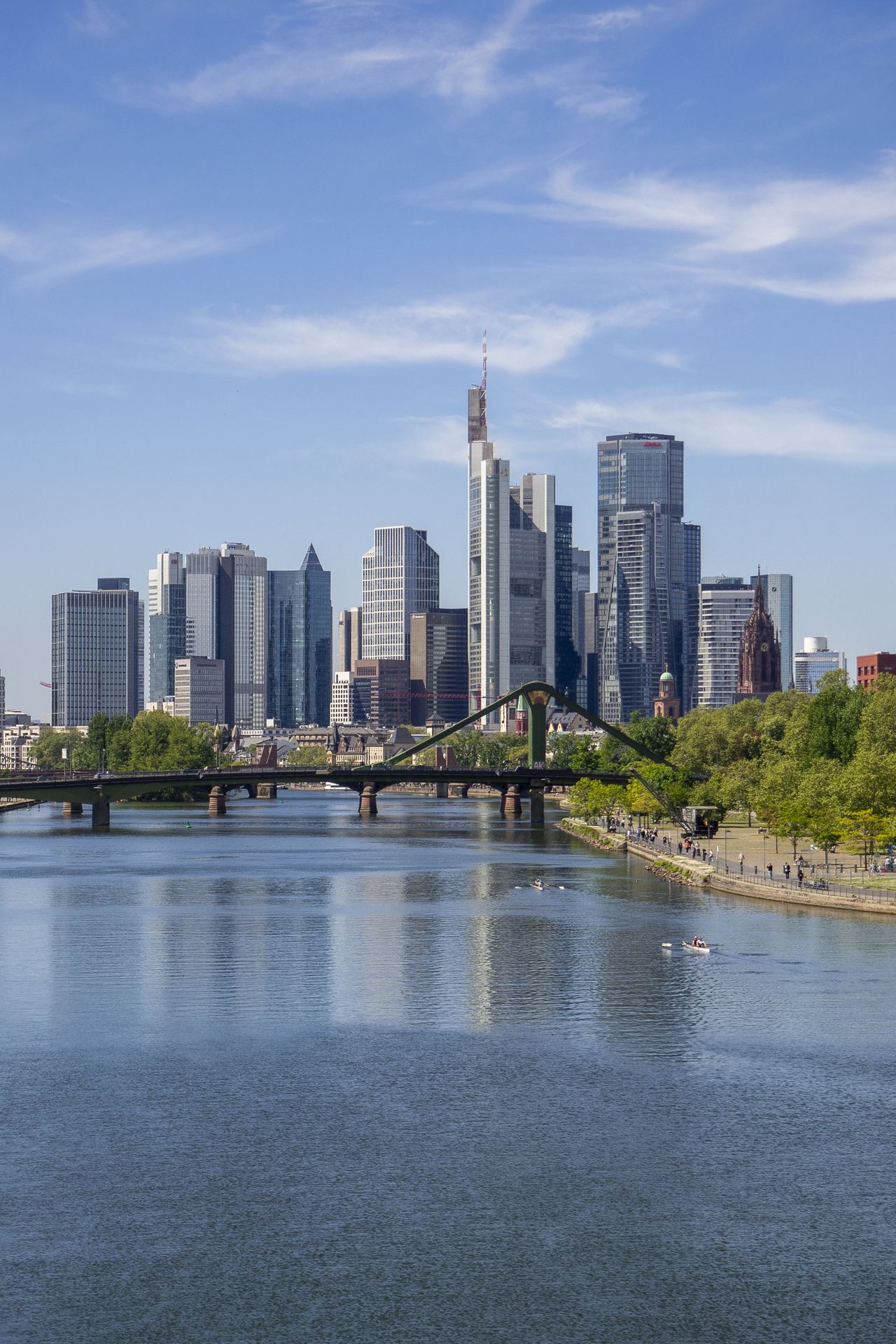 Blick auf die moderne Skyline einer Stadt mit hohen Glasgebäuden, einem Fluss im Vordergrund und einer grünen Uferpromenade bei klarem Himmel.