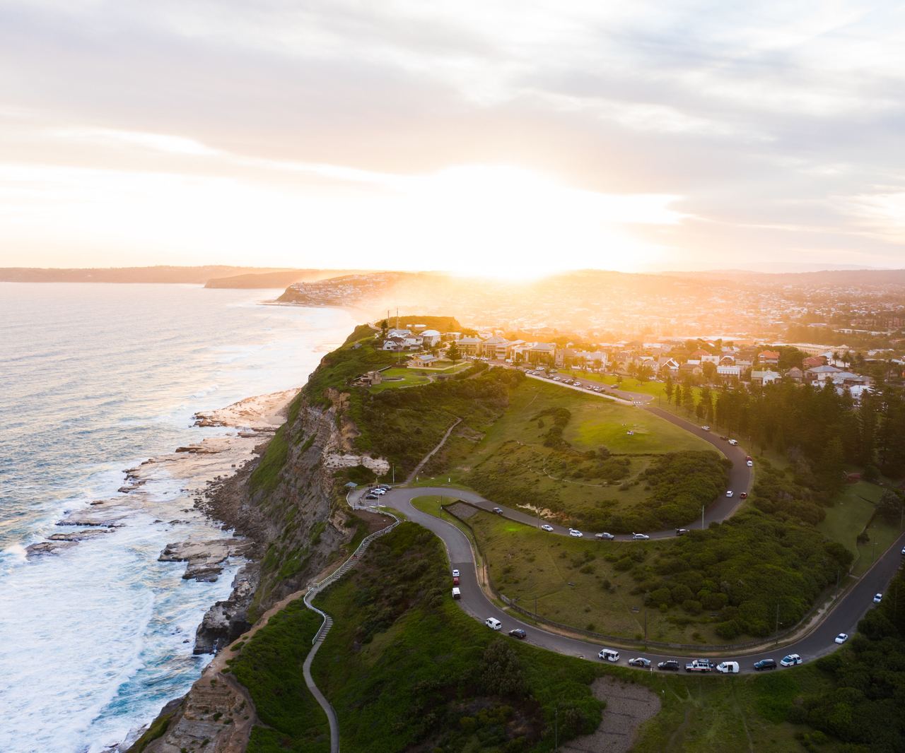 Luftaufnahme einer Küstenlandschaft bei Sonnenuntergang mit einer kurvigen Straße auf einer grünen Klippe und Blick auf das Meer sowie einen Ort im Hintergrund. Die Szene vermittelt Ruhe und Weite.