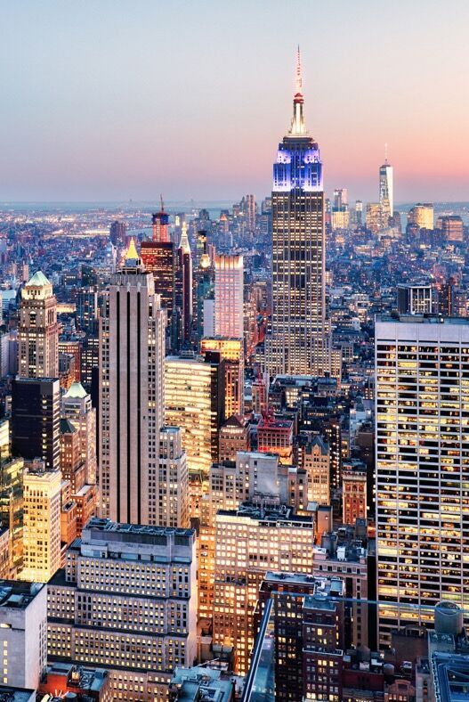 Blick auf die Skyline von New York bei Dämmerung mit dem beleuchteten Empire State Building im Zentrum, umgeben von hoch aufragenden Wolkenkratzern und der Stadt, die sich bis zum Horizont erstreckt.