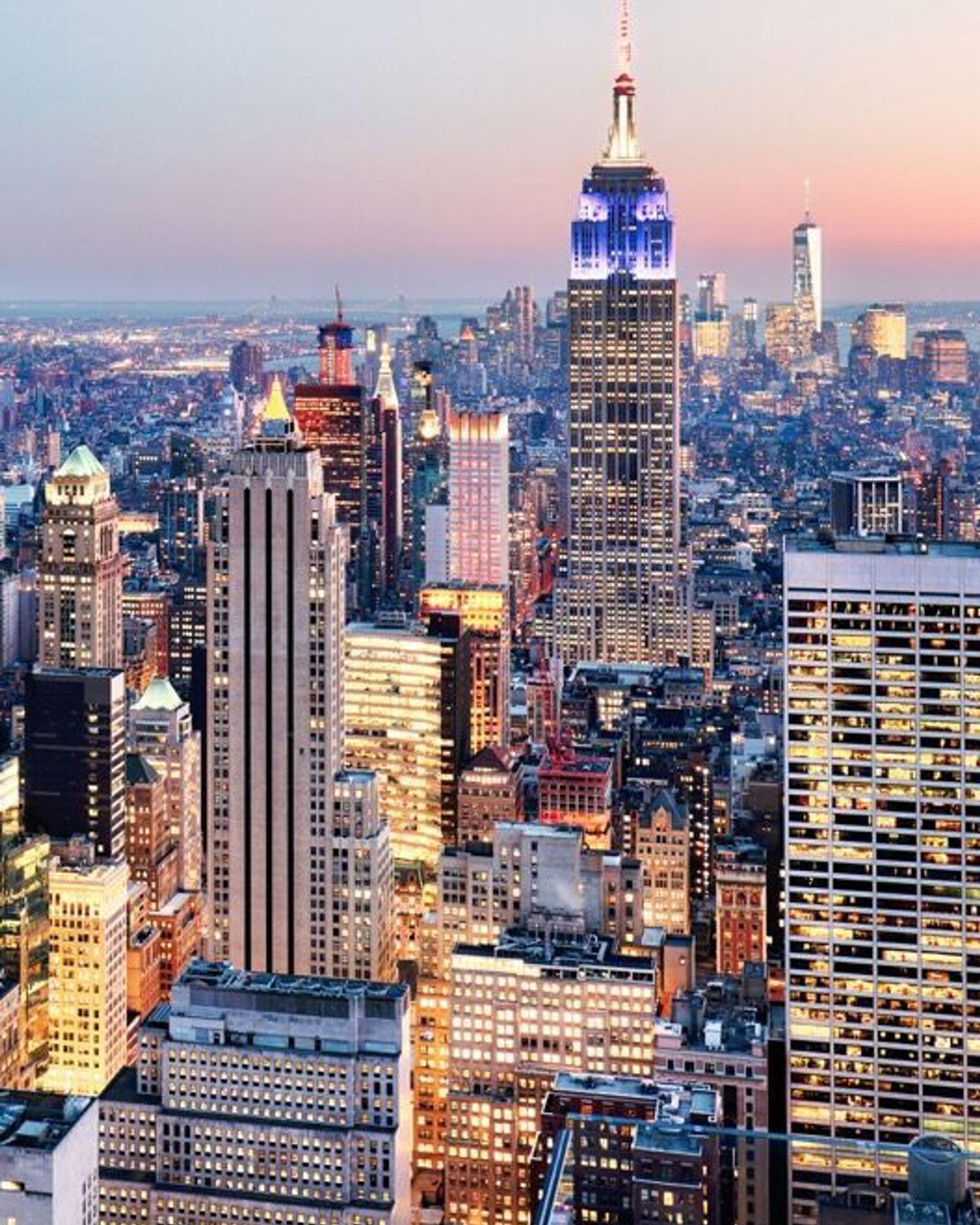 Blick auf die Skyline von New York bei Dämmerung mit dem beleuchteten Empire State Building im Zentrum, umgeben von hoch aufragenden Wolkenkratzern und der Stadt, die sich bis zum Horizont erstreckt.