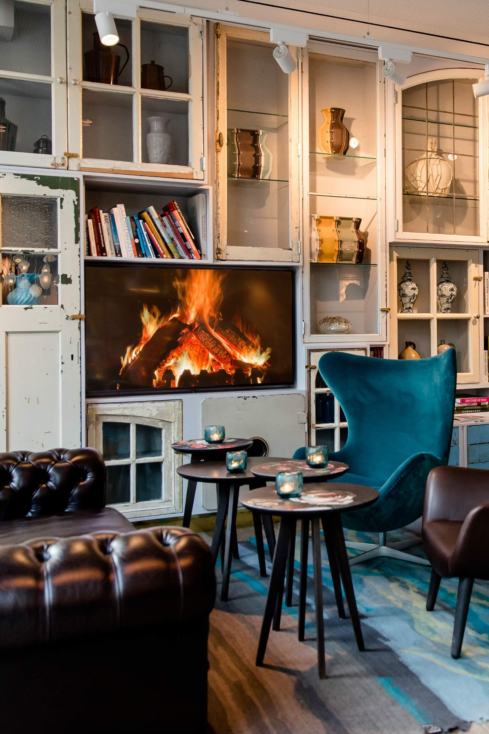 Gemütlicher Lounge-Bereich mit Ledersofa, Sesseln und kleinem Tisch vor einem Kaminfeuer, umgeben von Regalen mit Dekoration und Büchern.