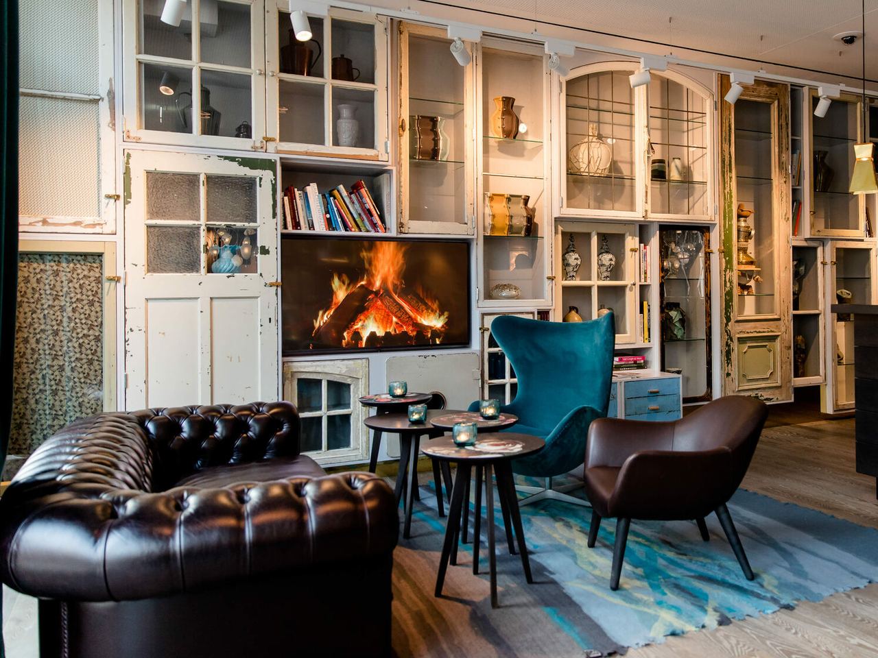 Gemütlicher Lounge-Bereich mit Ledersofa, Sesseln und kleinem Tisch vor einem Kaminfeuer, umgeben von Regalen mit Dekoration und Büchern.