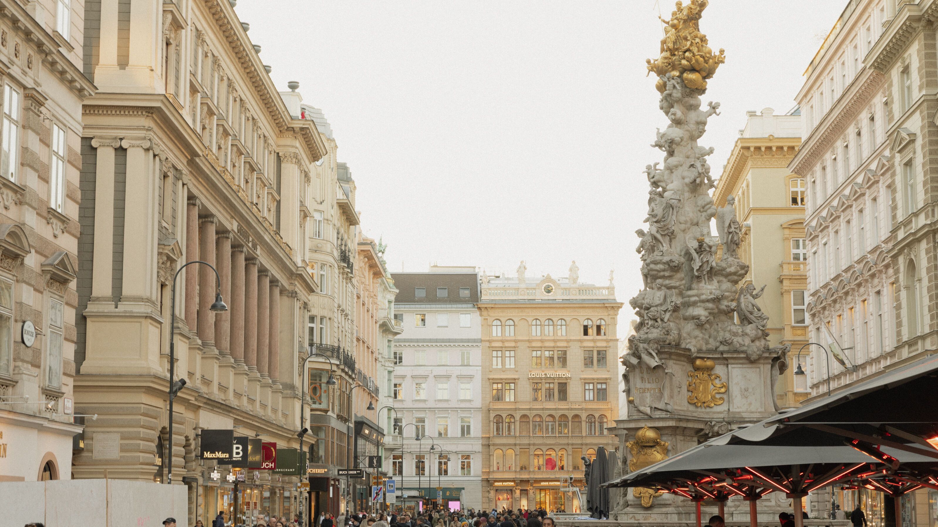 Belebte Fußgängerzone in einer historischen Stadt mit eleganten Gebäuden, vielen Passanten und einem kunstvollen Denkmal im Hintergrund. Außenbereiche von Cafés laden zum Verweilen ein.