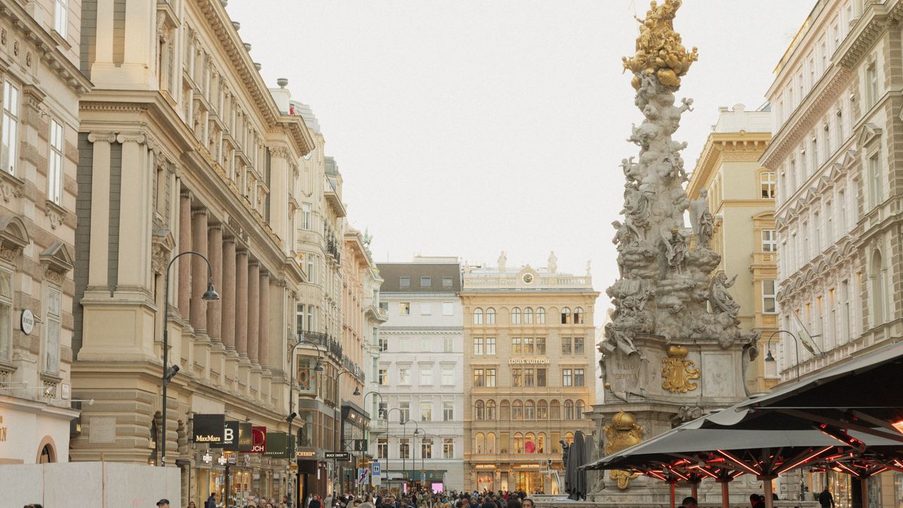 Belebte Fußgängerzone in einer historischen Stadt mit eleganten Gebäuden, vielen Passanten und einem kunstvollen Denkmal im Hintergrund. Außenbereiche von Cafés laden zum Verweilen ein.