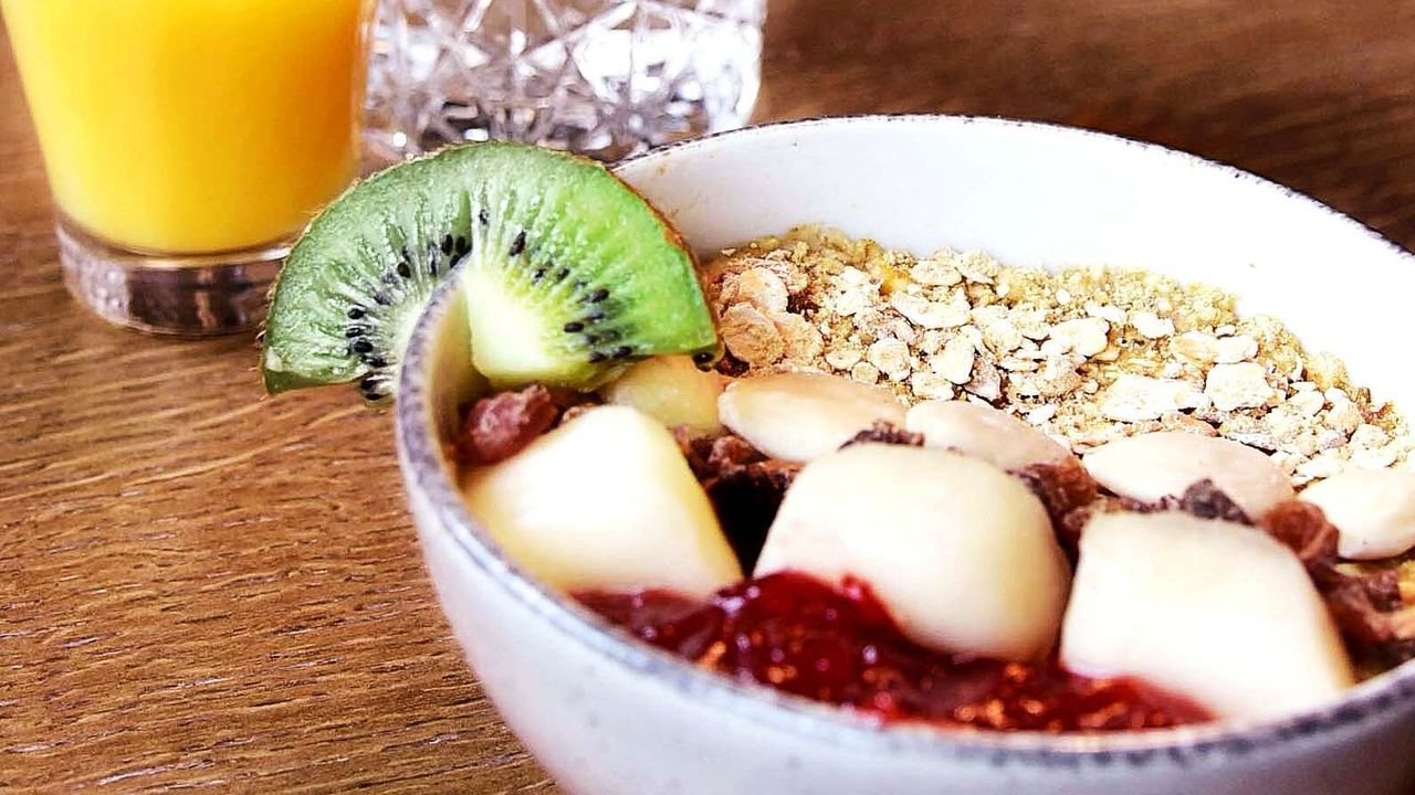 Schale mit Müsli, frischen Früchten und Nüssen, garniert mit einer Kiwischeibe, dazu ein Glas Orangensaft auf einem Holztisch.