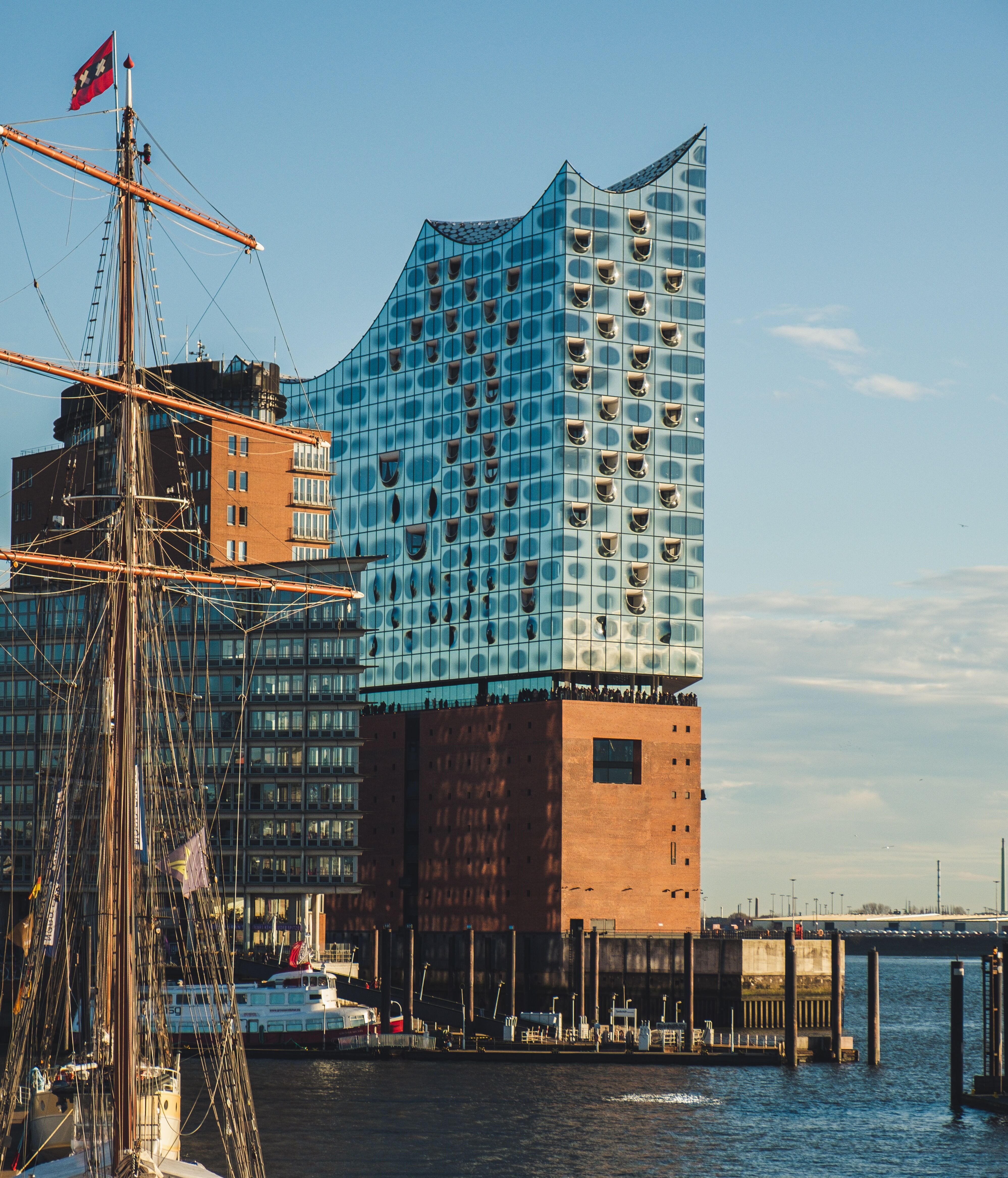 Modernes Gebäude mit wellenförmiger Glasfassade am Wasser, vor einem klaren Himmel, neben Segelschiffen im Hafen.